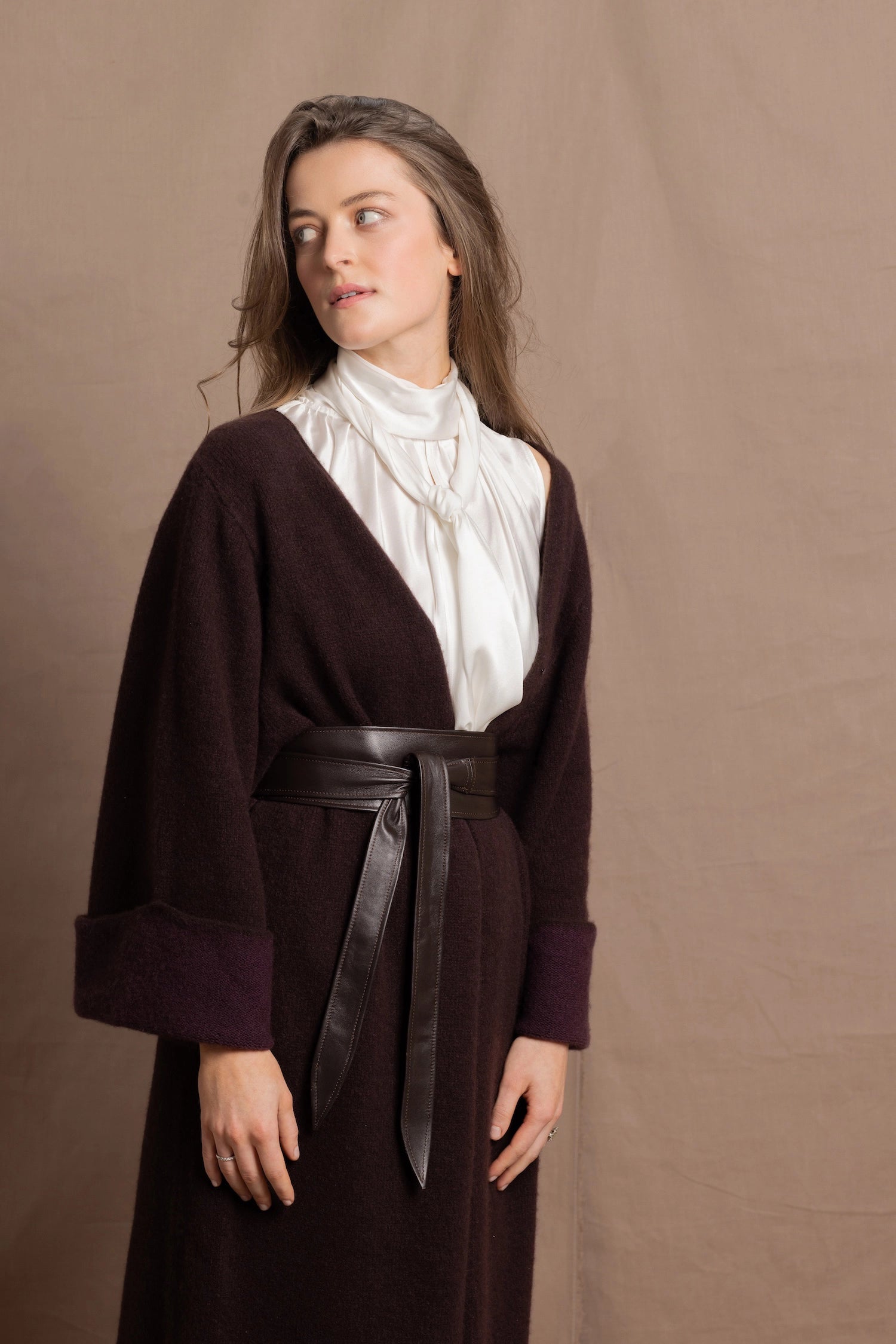 A woman wears a 100% silk pussybow sleeveless blouse in white with a chocolate brown and purple cashmere coat on top and cinched with leather obi belt