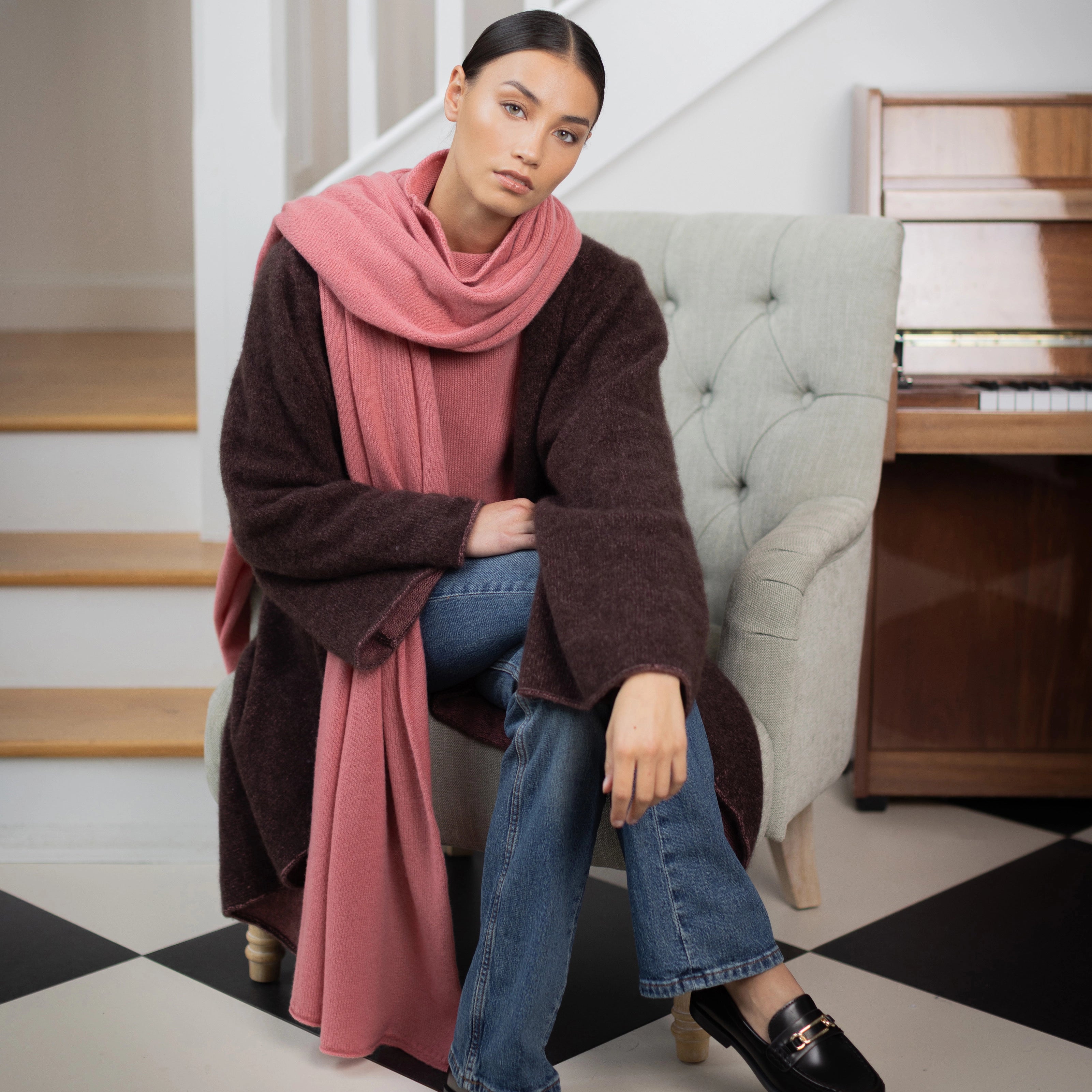 a woman wears a luxurious cashmere wrap in a dusty rose colour with matching cashmere sweater and a dark brown cashmere coat over blue jeans with brown loafers.