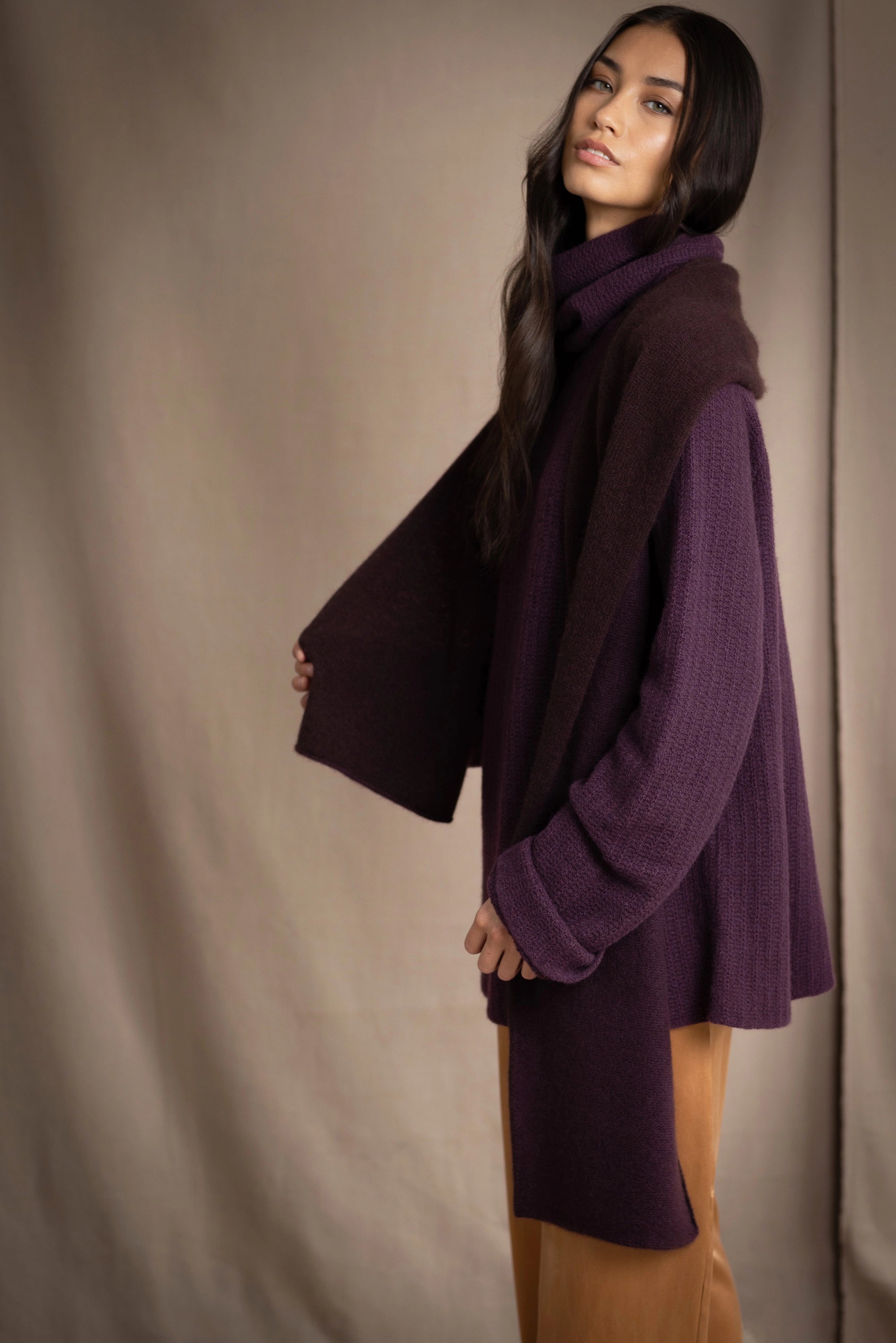 woman wears a reversible cashmere scarf in dark purple and dark brown on the other side worn over a dark purple cashmere sweater and bronze silk trousers