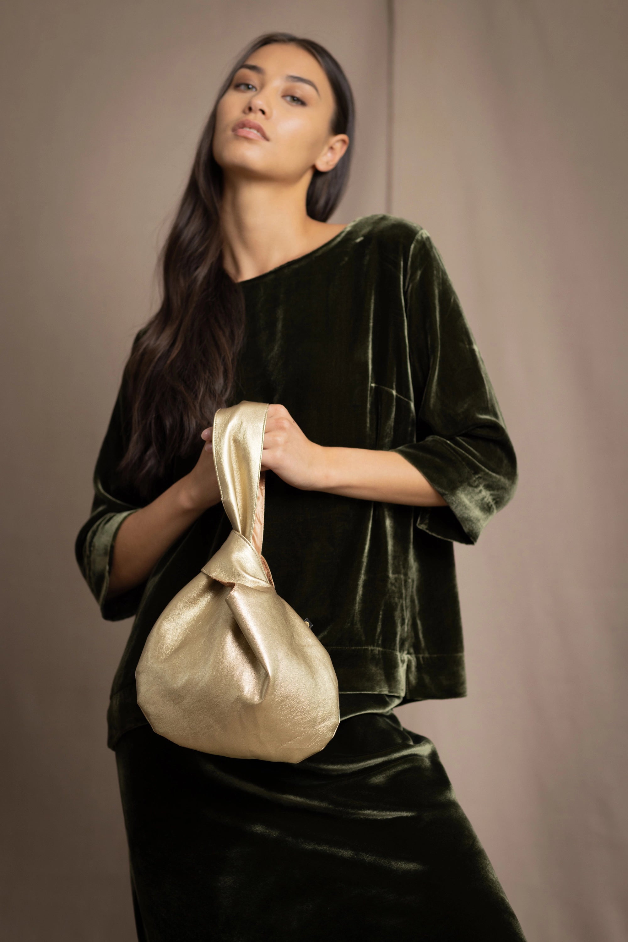 Gold vegan leather pull through handbag held by a person wearing dark green silk velvet clothing