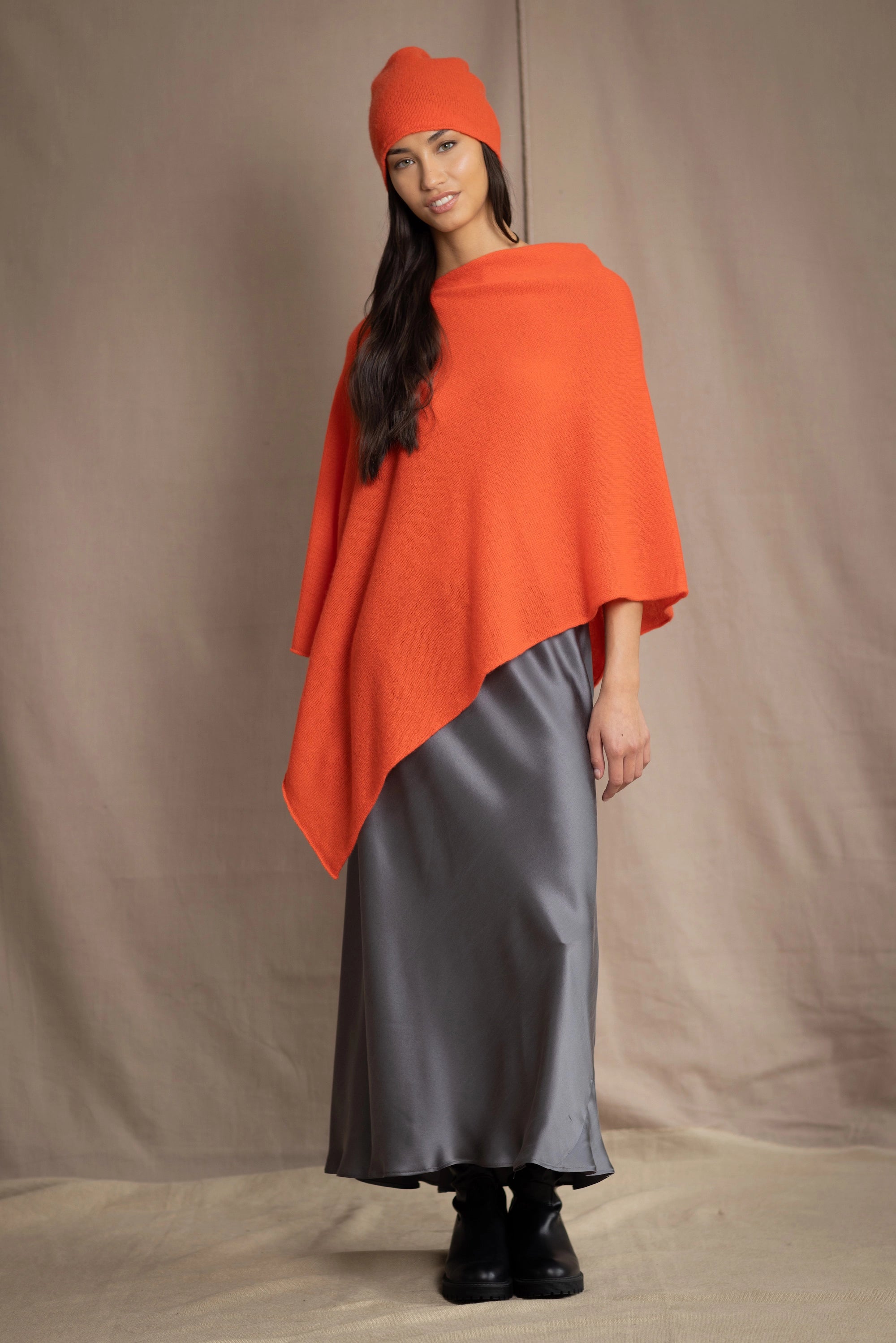 a woman wears a cashmere cape in orange with matching cashmere beanie hat over grey silk skirt