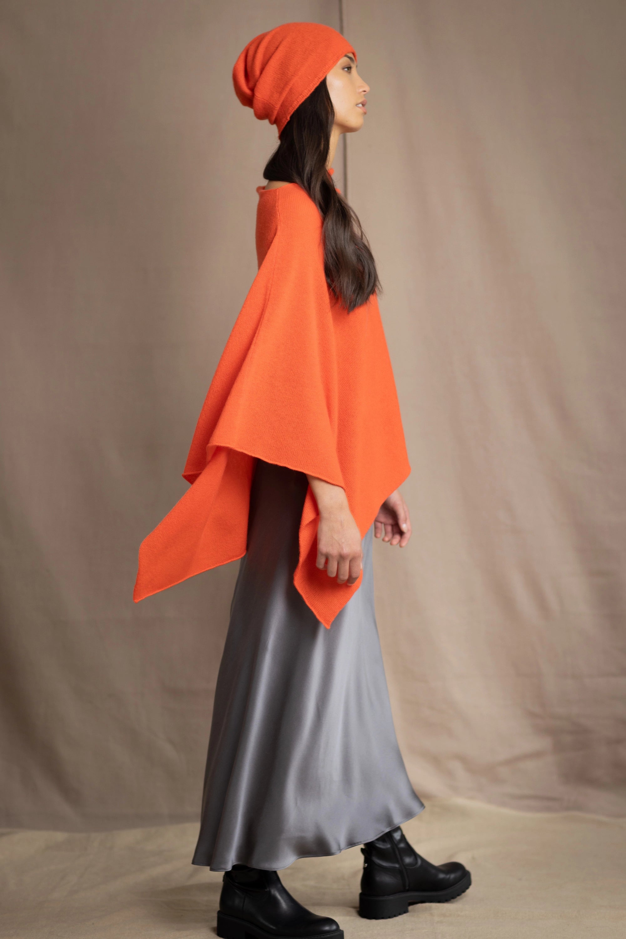 a woman wears a cashmere cape in orange with matching cashmere beanie hat over grey silk skirt