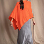 a woman wears a cashmere cape in orange with matching cashmere beanie hat over grey silk skirt