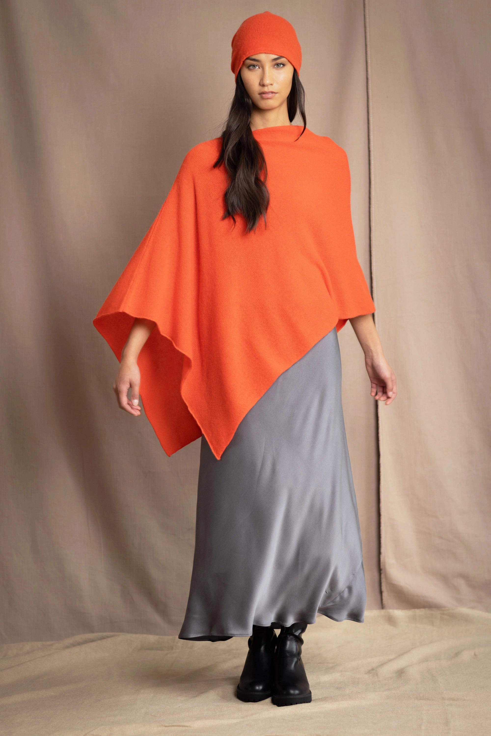 a woman wears a cashmere cape in orange with matching cashmere beanie hat over grey silk skirt