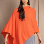 a woman wears a cashmere cape in orange with matching cashmere beanie hat over grey silk skirt