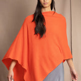 a woman wears a cashmere cape in orange with matching cashmere beanie hat over grey silk skirt