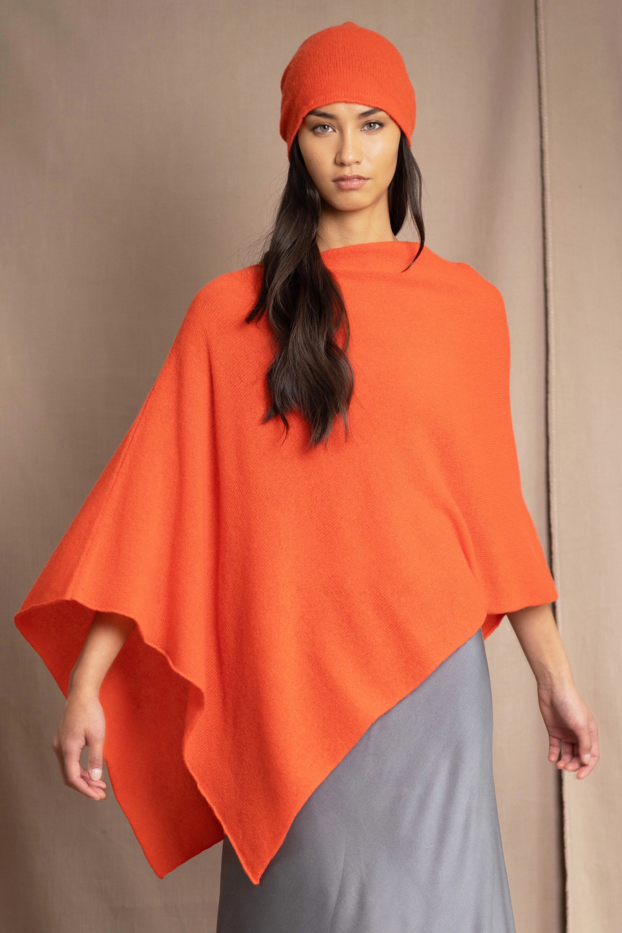 a woman wears a cashmere cape in orange with matching cashmere beanie hat over grey silk skirt