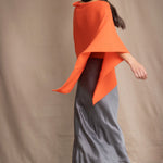 a woman wears a cashmere cape in orange with matching cashmere beanie hat over grey silk skirt