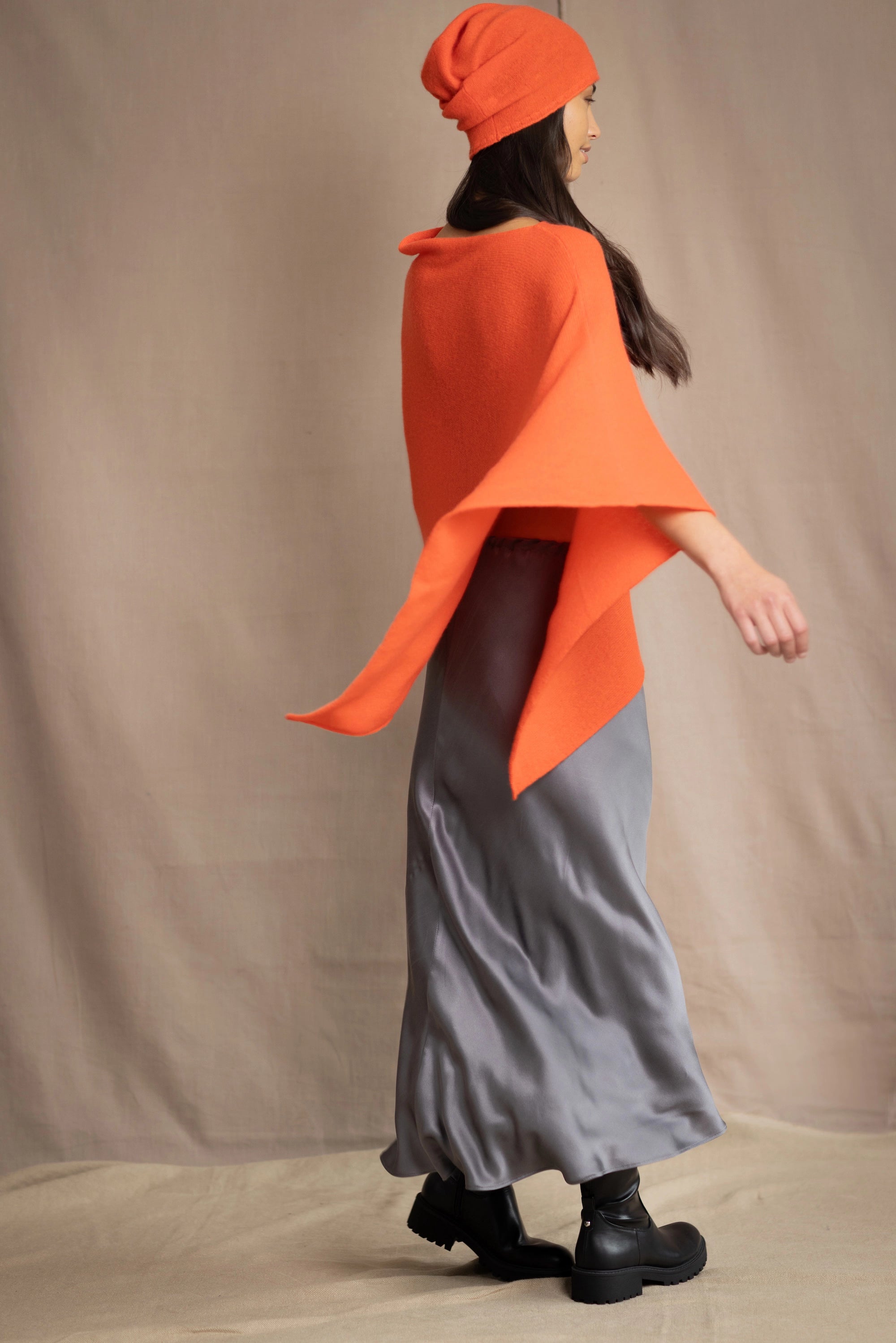 a woman wears a cashmere cape in orange with matching cashmere beanie hat over grey silk skirt