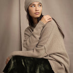a woman wearing an oversized cashmere sweater in beige with gold metallic lurex thread and matching cashmere beanie hat over a silk velvet skirt in green