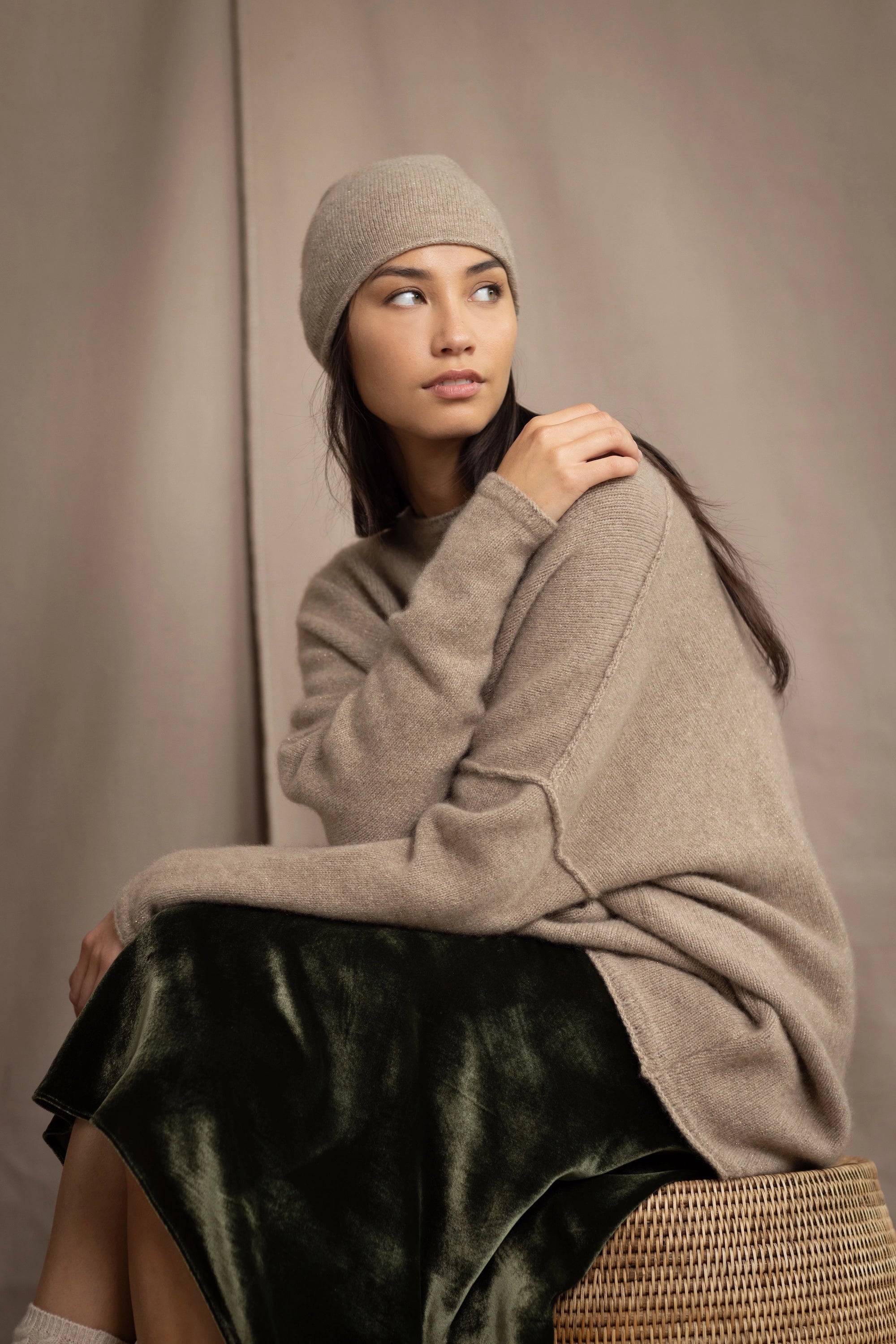a woman wearing an oversized cashmere sweater in beige with gold metallic lurex thread and matching cashmere beanie hat over a silk velvet skirt in green