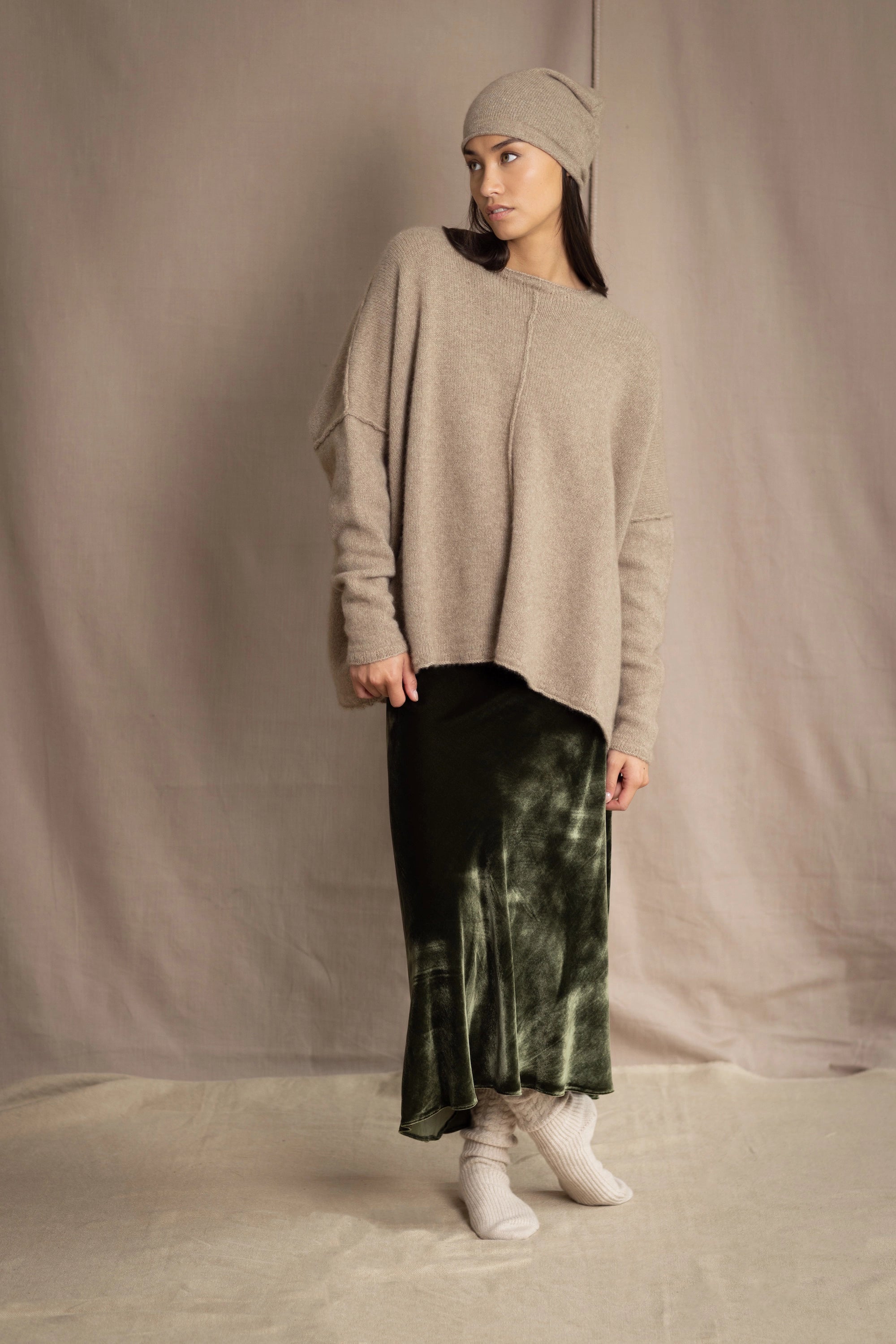 a woman wearing an oversized cashmere sweater in beige with gold metallic lurex thread and matching cashmere beanie hat over a silk velvet skirt in green
