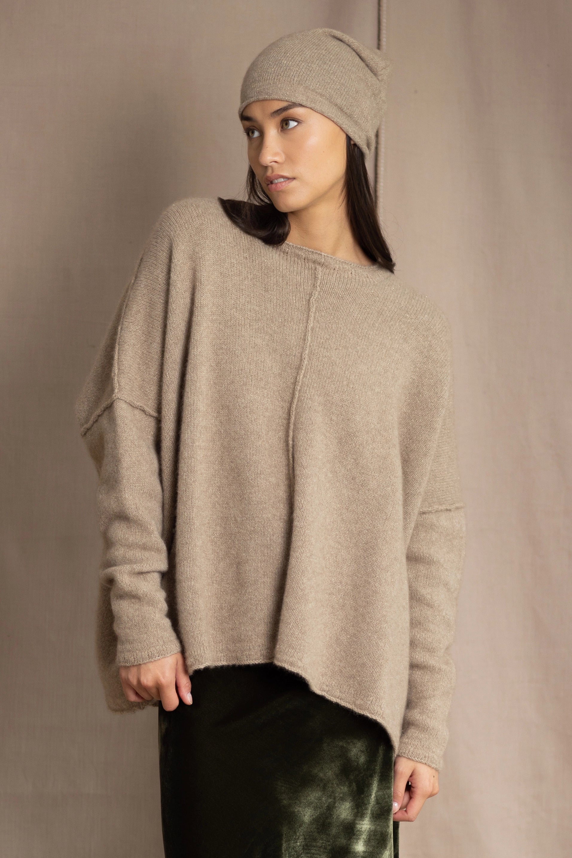 a woman wearing an oversized cashmere sweater in beige with gold metallic lurex thread and matching cashmere beanie hat over a silk velvet skirt in green