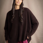 a woman wearing an oversized cashmere sweater in dark brown with matching cashmere beanie hat over silk trousers in burgundy