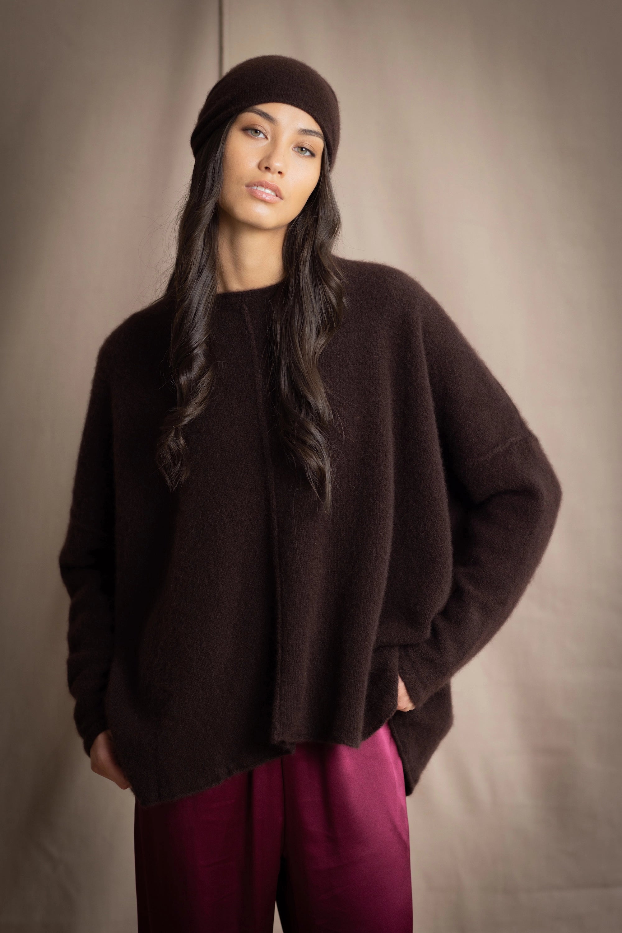 a woman wearing an oversized cashmere sweater in dark brown with matching cashmere beanie hat over silk trousers in burgundy
