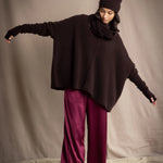 a woman wearing an oversized cashmere sweater in dark brown with matching cashmere beanie hat, snood and fingerless gloves over silk trousers in burgundy