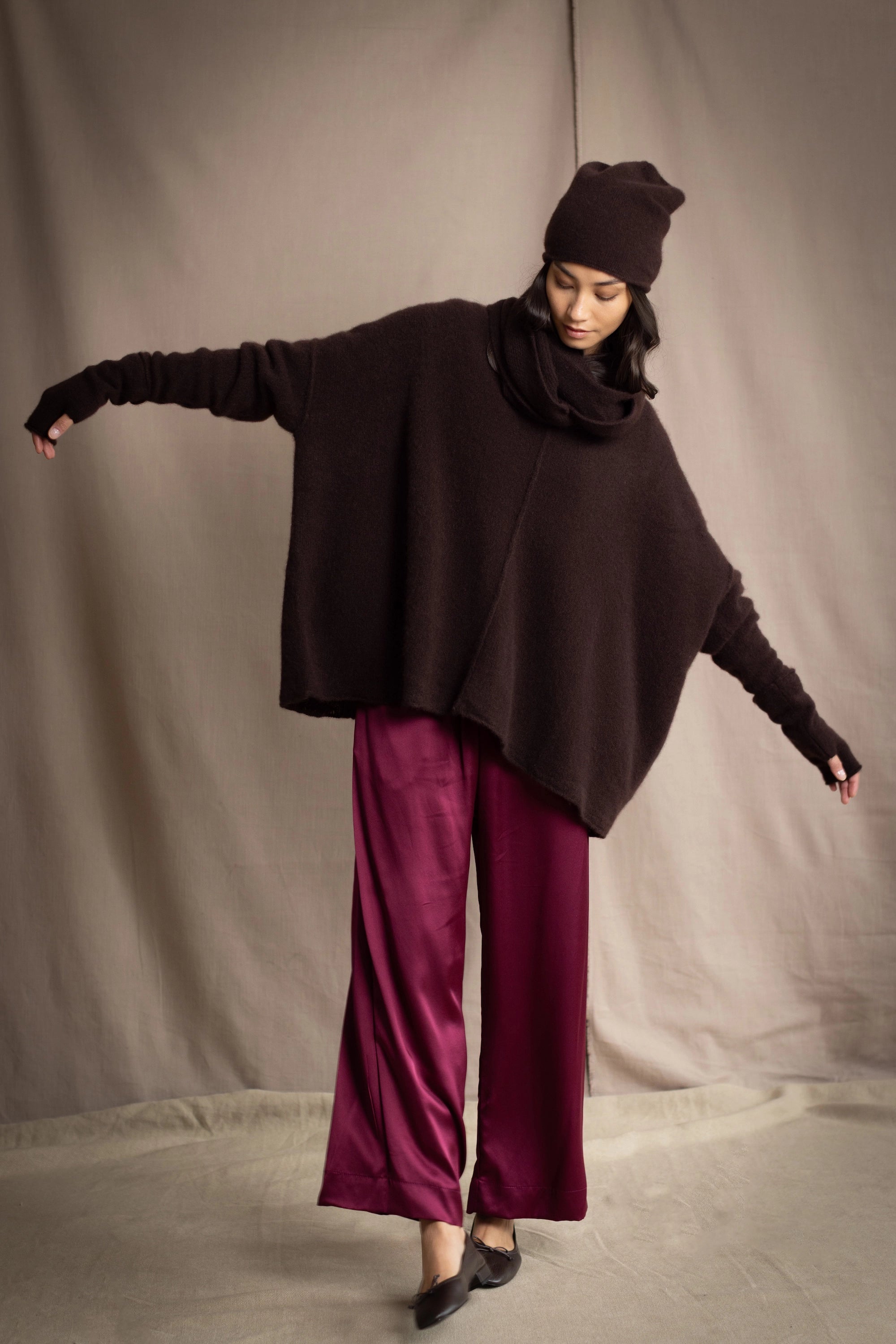 a woman wearing an oversized cashmere sweater in dark brown with matching cashmere beanie hat, snood and fingerless gloves over silk trousers in burgundy