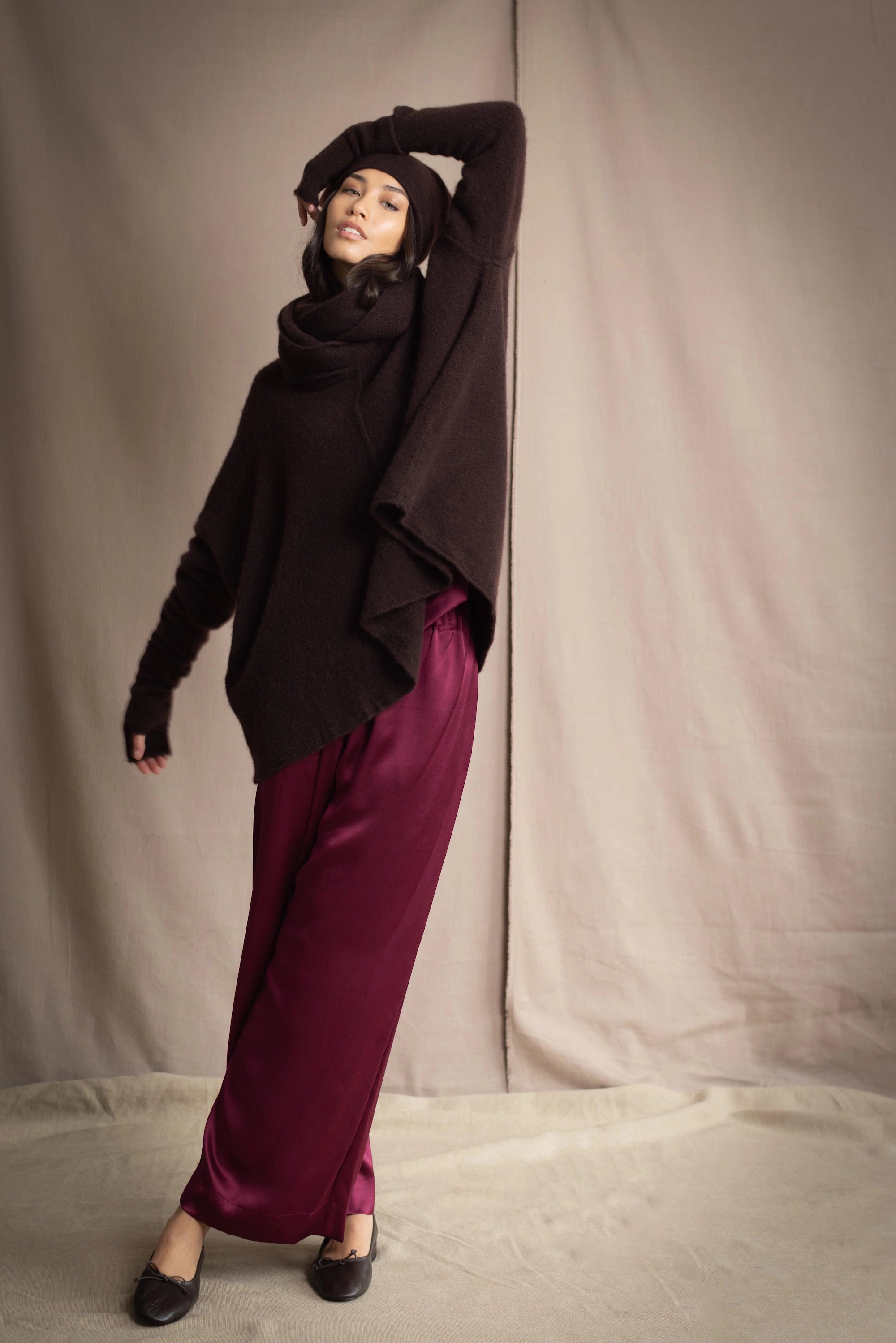 a woman wearing an oversized cashmere sweater in dark brown with matching cashmere beanie hat, snood and fingerless gloves over silk trousers in burgundy