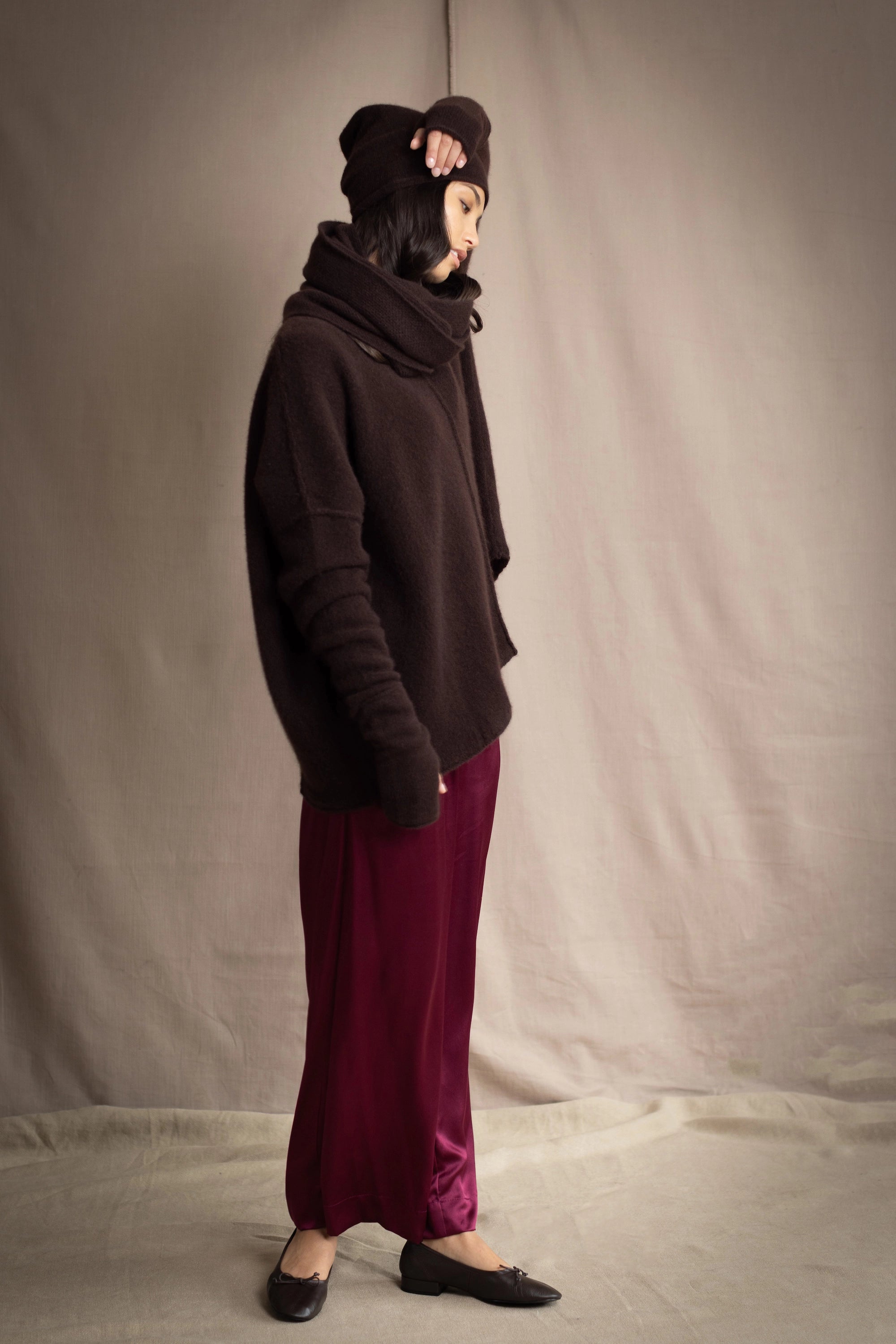 a woman wearing an oversized cashmere sweater in dark brown with matching cashmere beanie hat, snood and fingerless gloves over silk trousers in burgundy