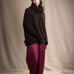a woman wearing an oversized cashmere sweater in dark brown with matching cashmere beanie hat, snood and fingerless gloves over silk trousers in burgundy