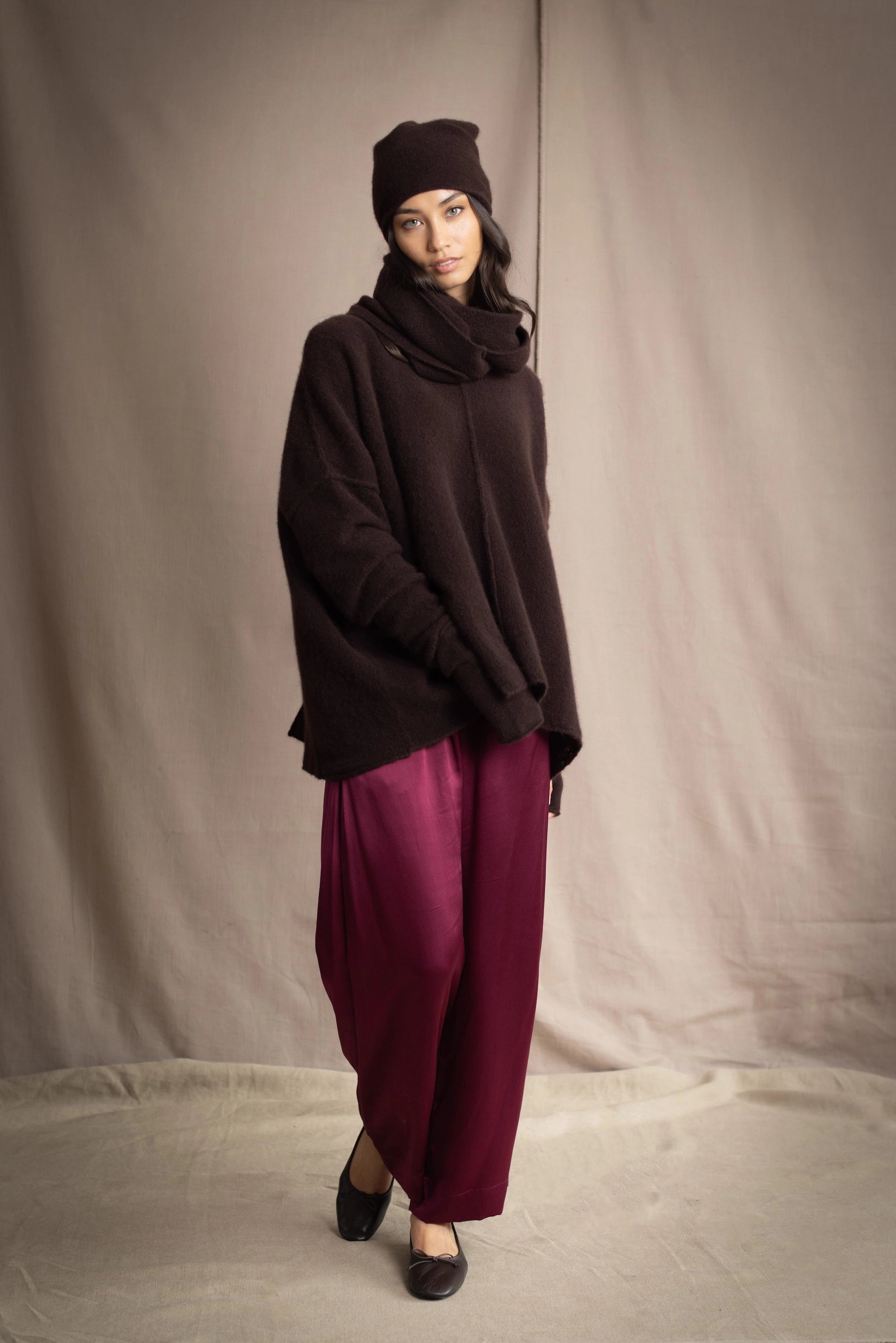a woman wearing an oversized cashmere sweater in dark brown with matching cashmere beanie hat, snood and fingerless gloves over silk trousers in burgundy
