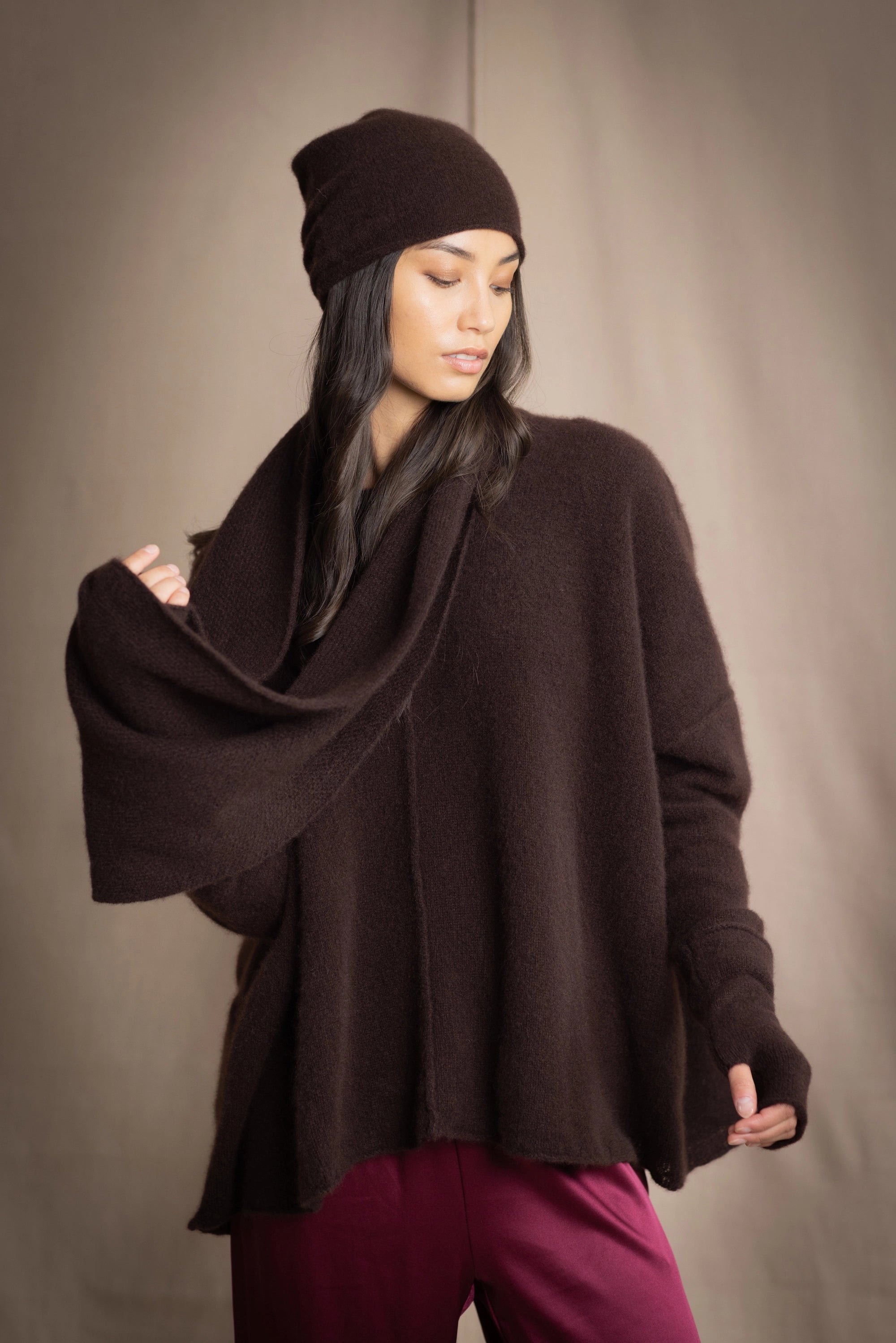 a woman wearing cashmere fingerless gloves in dark brown with matching cashmere beanie hat and cashmere snood and cashmere sweater with burgundy silk pants