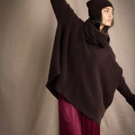 a woman wearing an oversized cashmere sweater in dark brown with matching cashmere beanie hat, snood and fingerless gloves over silk trousers in burgundy