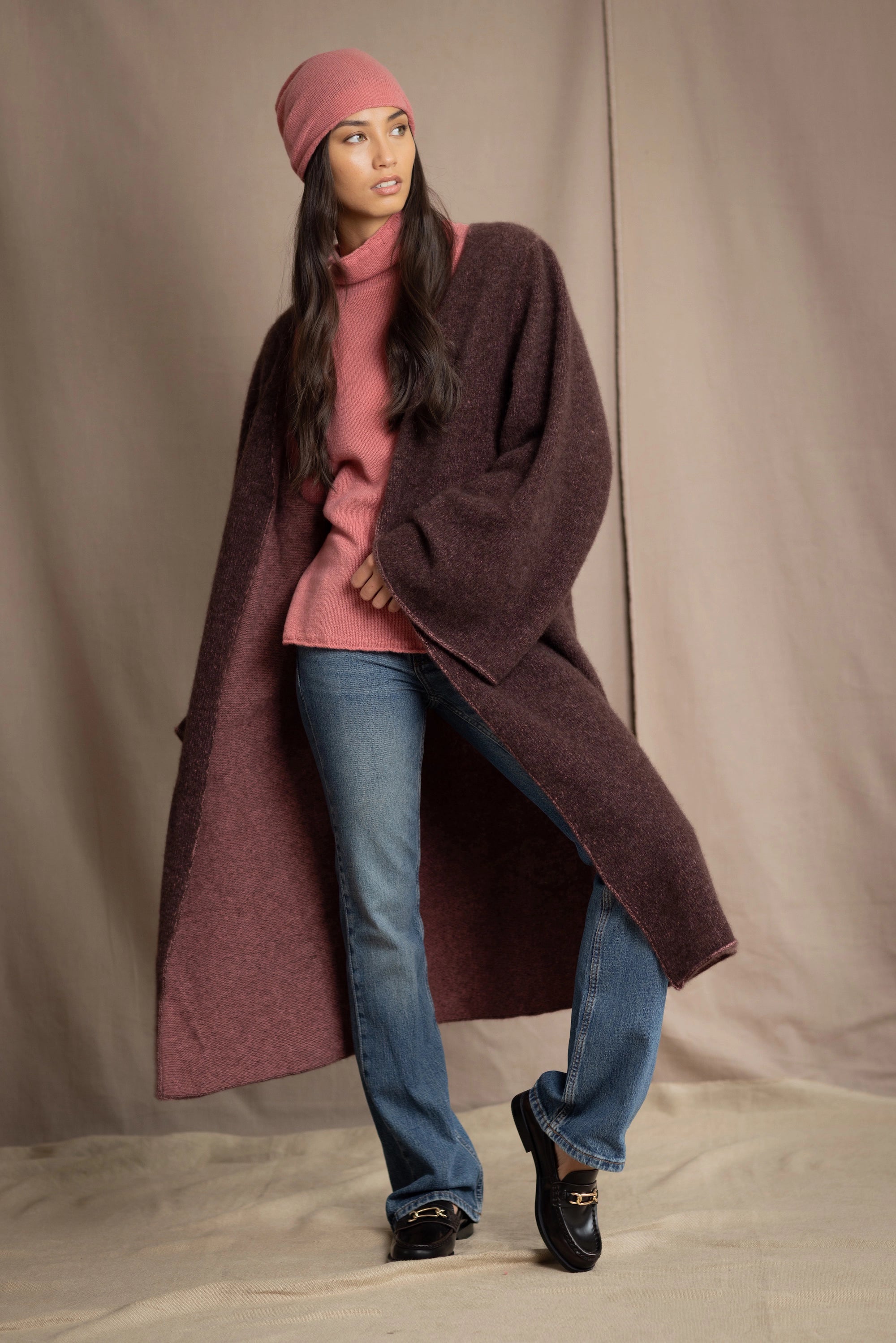 Woman wearing a long brown cashmere coat, pink cashmere sweater, blue jeans, and pink cashmere beanie
