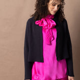 A woman wears a 100% cashmere cropped cardigan on top in navy blue over a 100% pure silk top in bright shocking pink over matching trousers