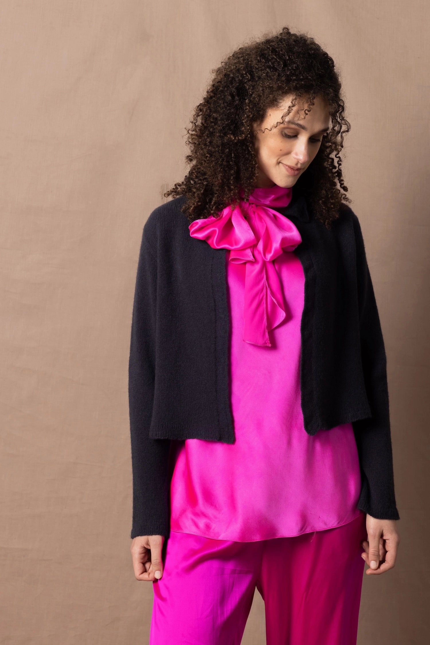 A woman wears a 100% cashmere cropped cardigan on top in navy blue over a 100% pure silk top in bright shocking pink over matching trousers