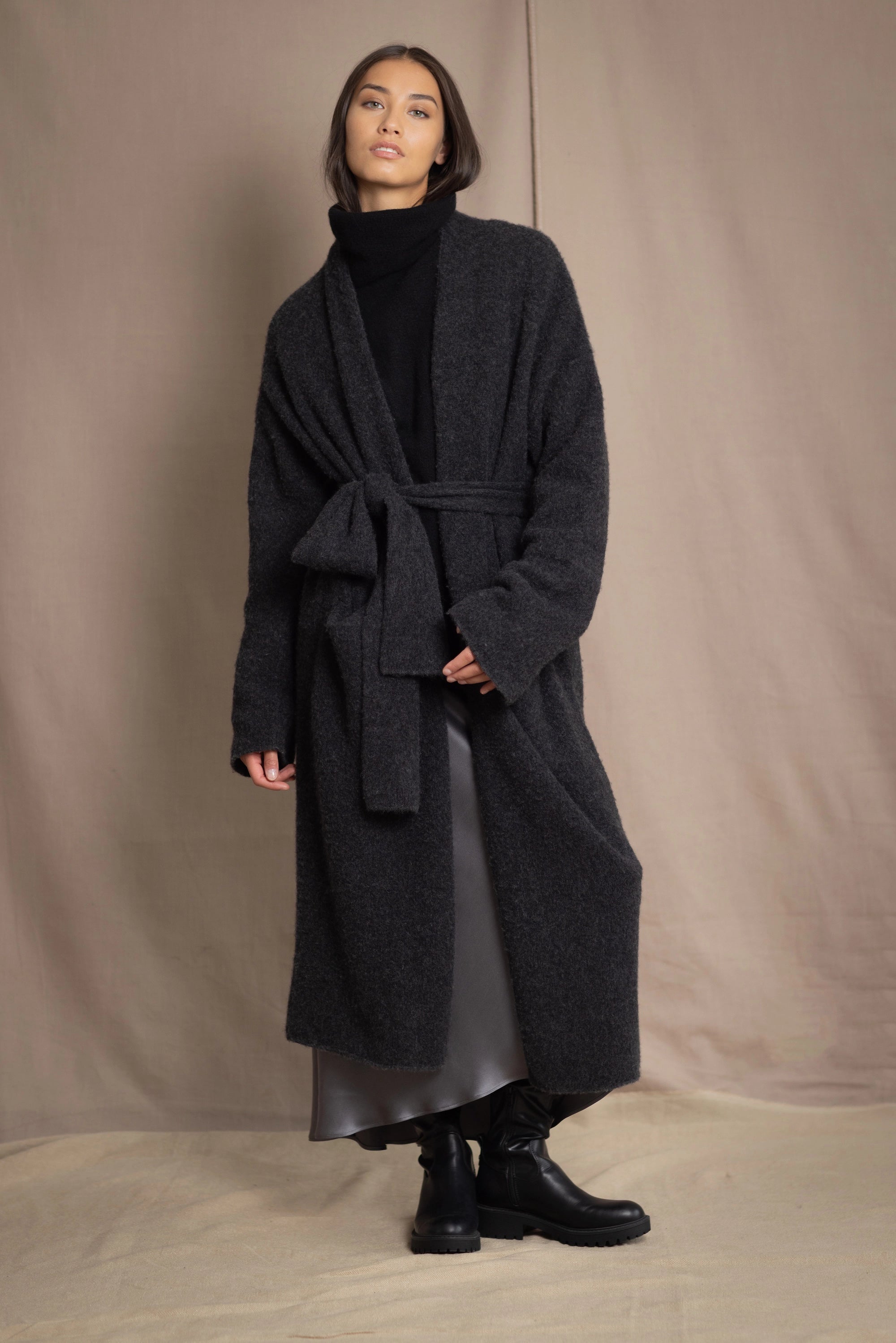 Woman wearing a long cashmere coat in dark grey over black cashmere turtleneck sweater and pewter silk skirt with black leather boots
