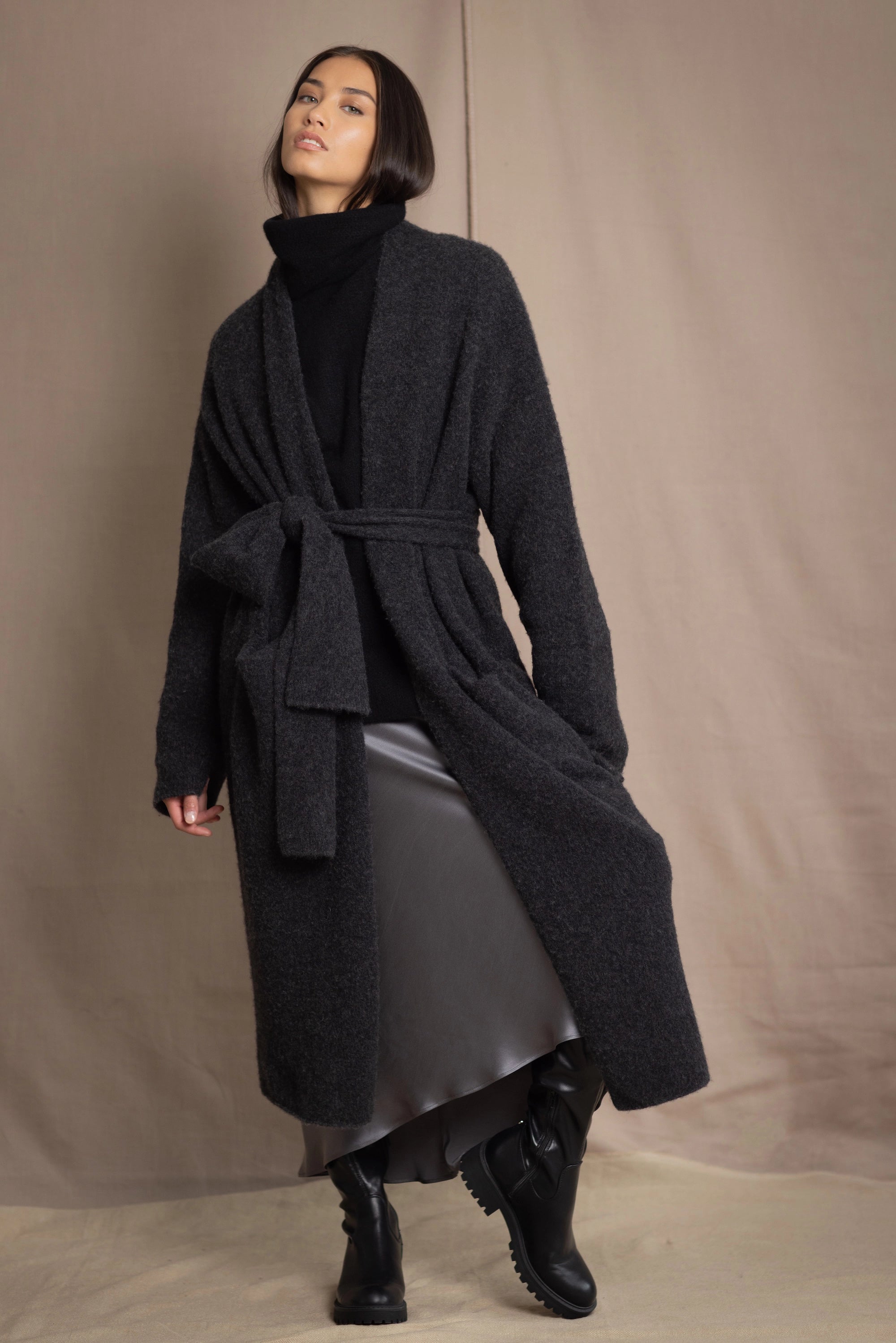 A woman wearing a long grey cashmere coat with black cashmere fisherman turtleneck sweater jumper with pewter silk skirt and black leather boots