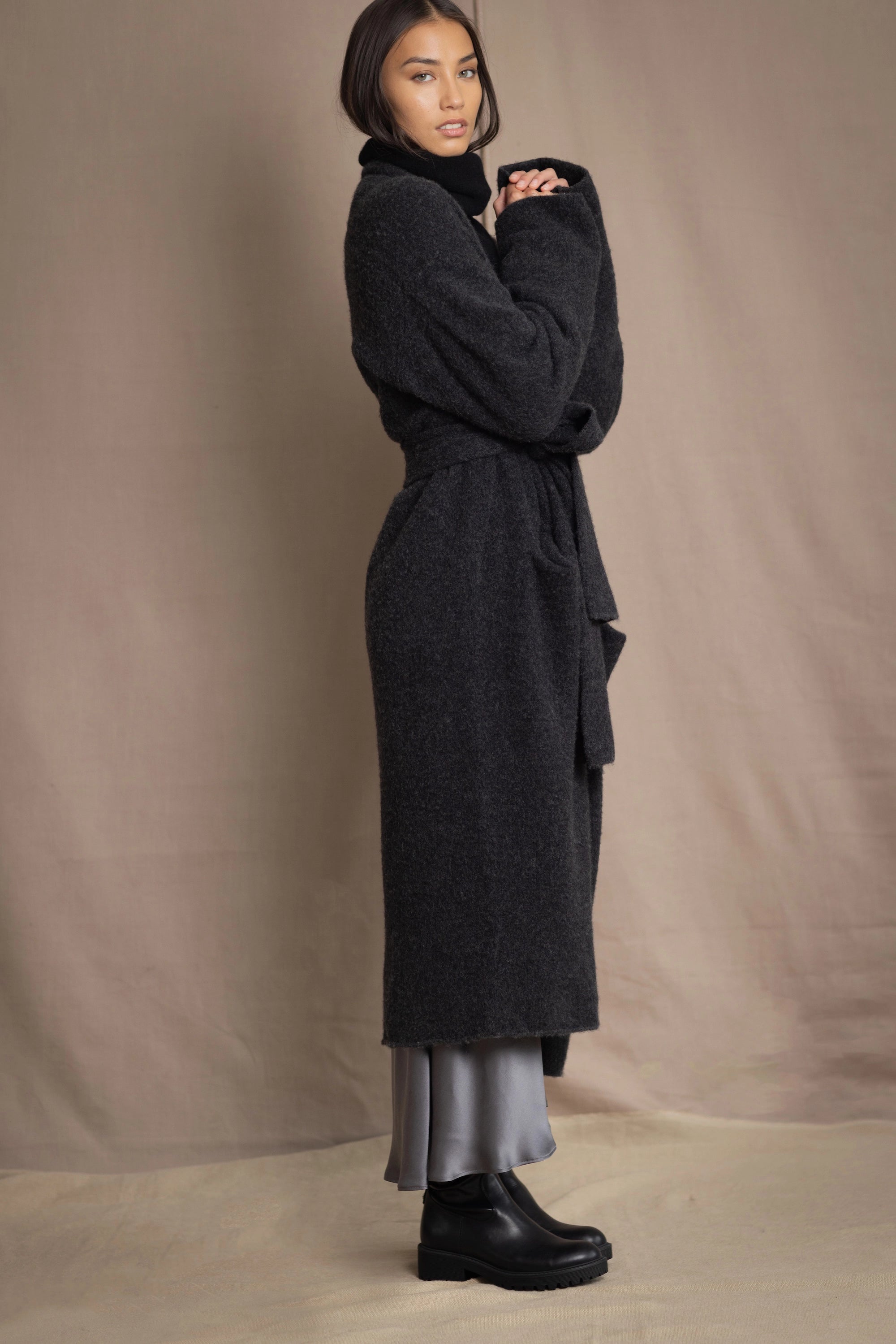 Woman wearing a long cashmere coat in dark grey over black cashmere turtleneck sweater and pewter silk skirt with black leather boots