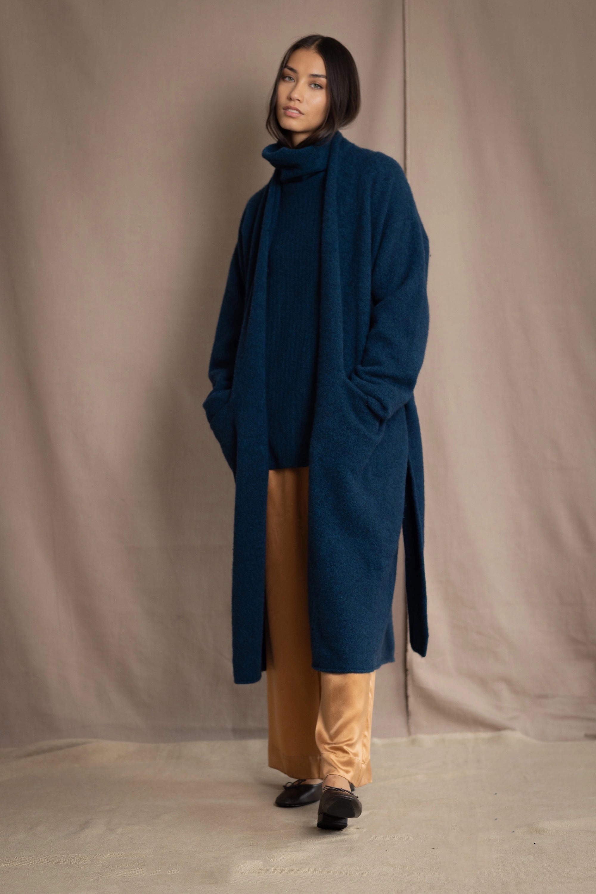 Woman wearing a long cashmere coat in teal blue over teal blue cashmere turtleneck sweater and bronze silk trousers