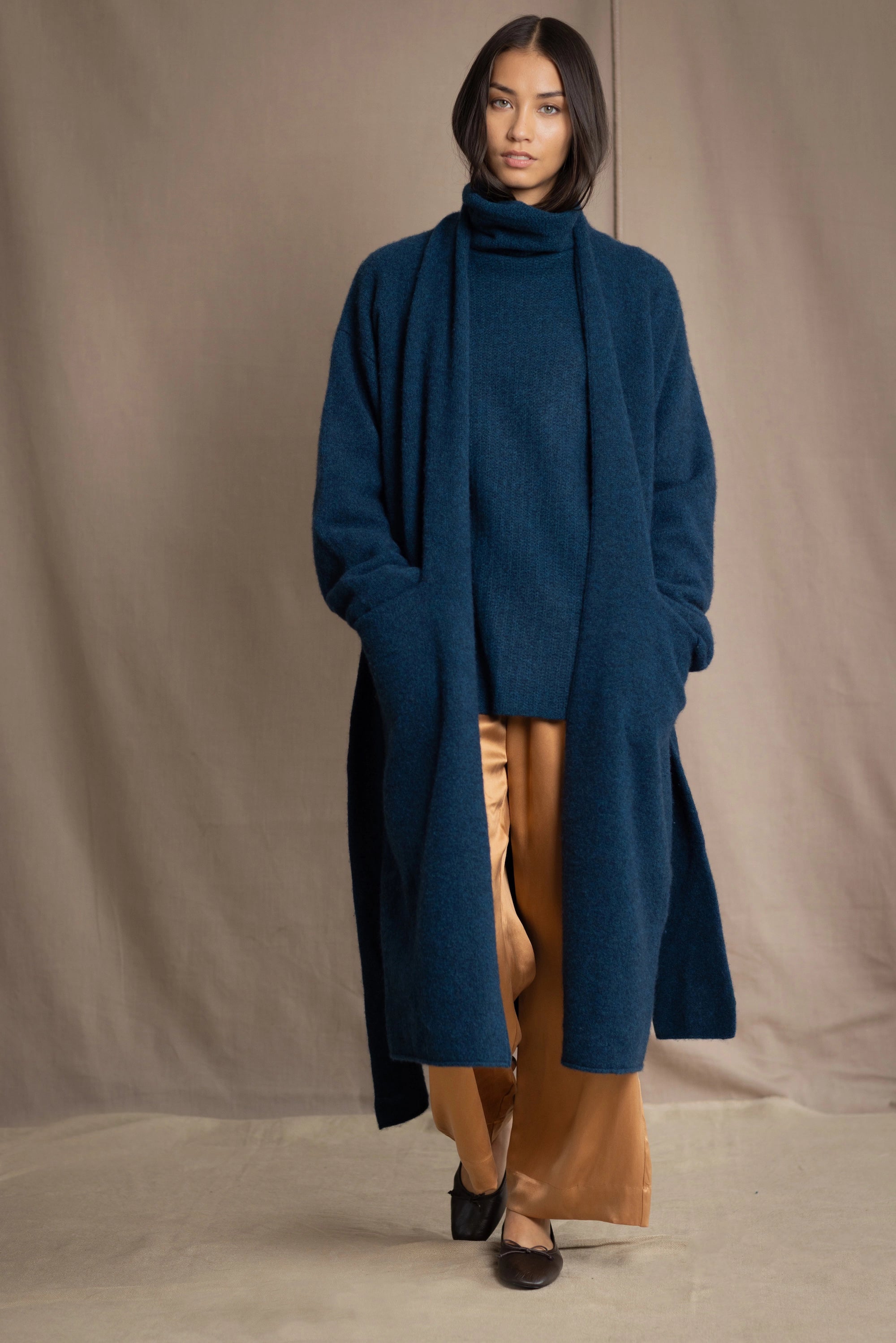 Woman wearing a long cashmere coat in teal blue over teal blue cashmere turtleneck sweater and bronze silk trousers