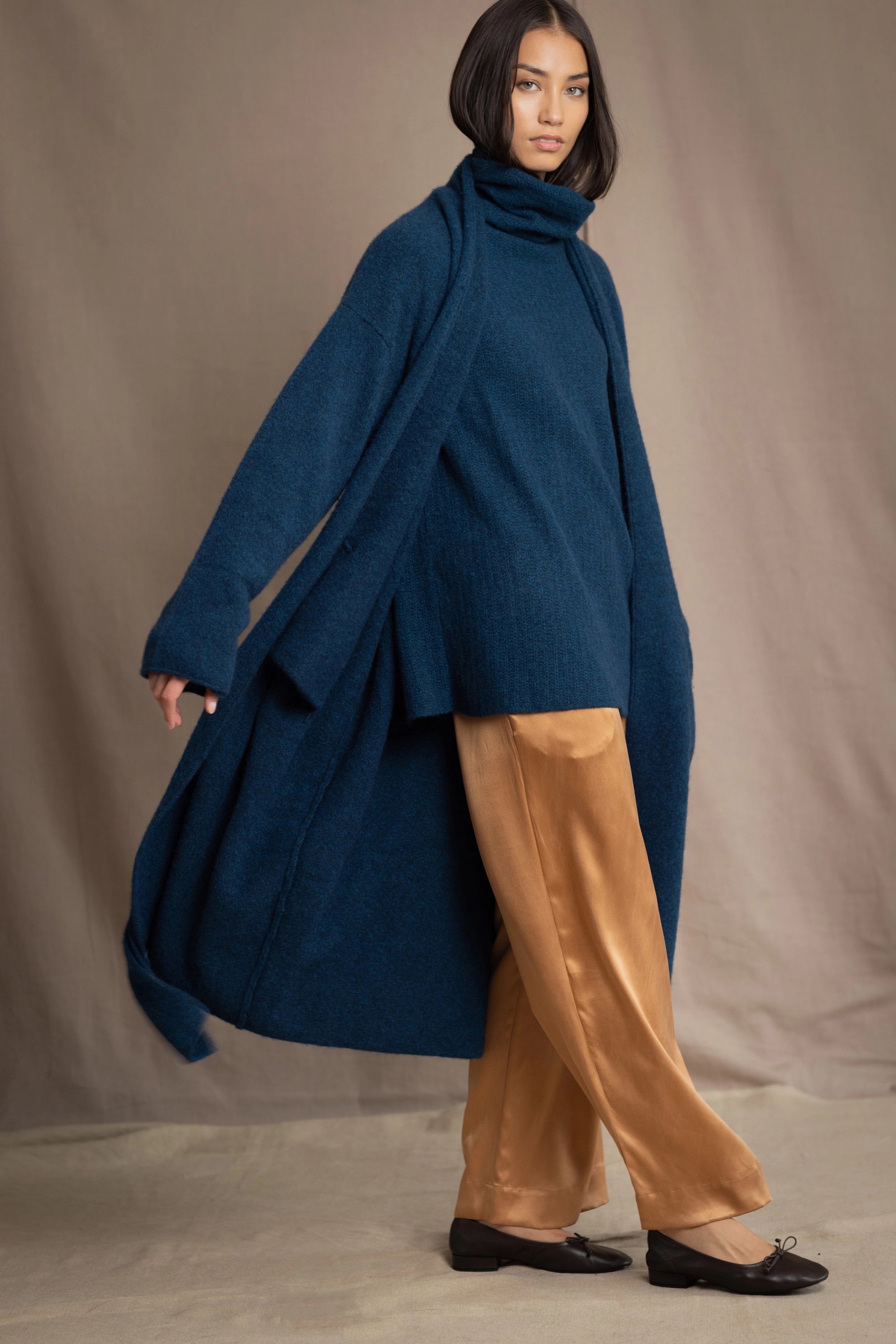 Woman wearing a long cashmere coat in teal blue over teal blue cashmere turtleneck sweater and bronze silk trousers