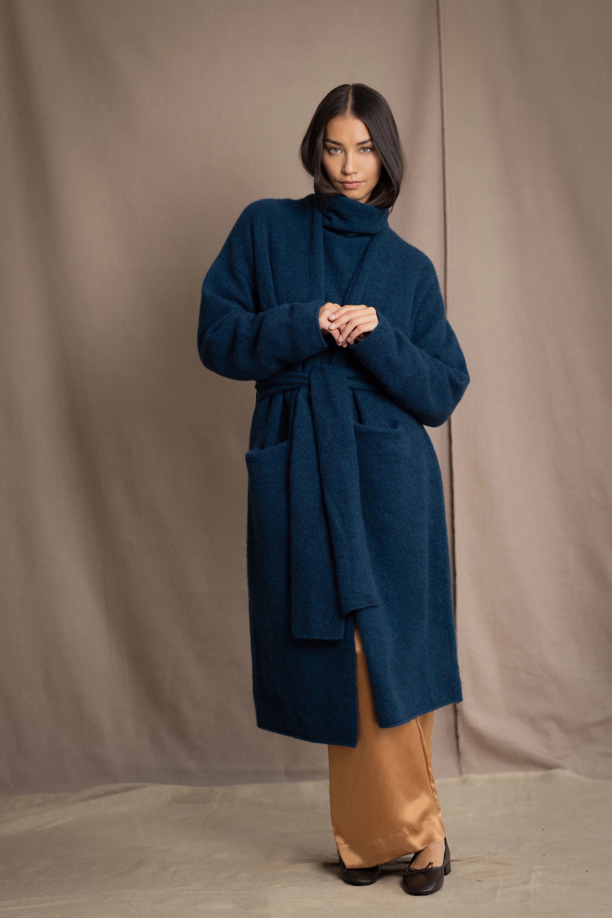 Woman wearing a long cashmere coat in teal blue over teal blue cashmere turtleneck sweater and bronze silk trousers