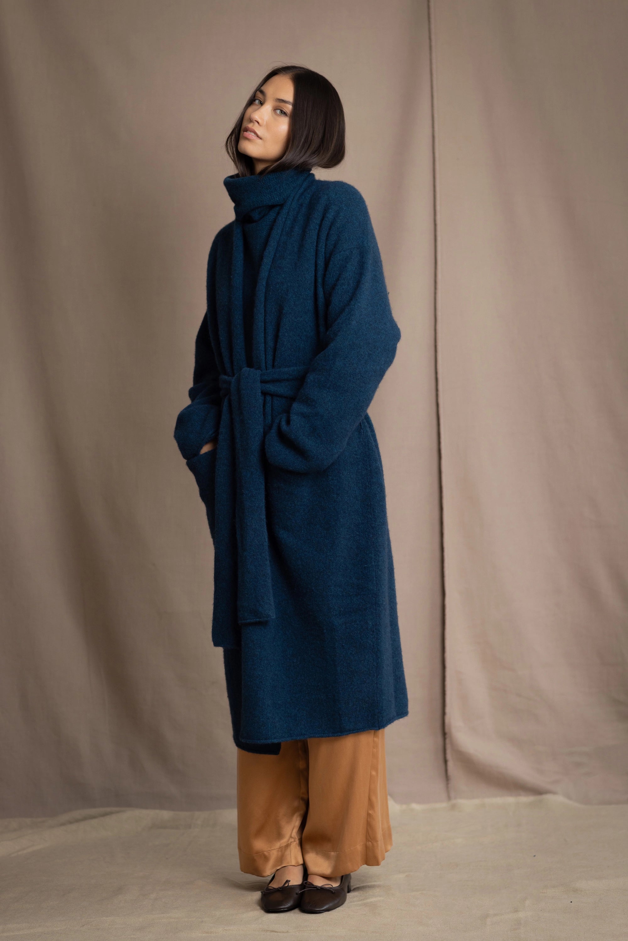 Woman wearing a long cashmere coat in teal blue over teal blue cashmere turtleneck sweater and bronze silk trousers