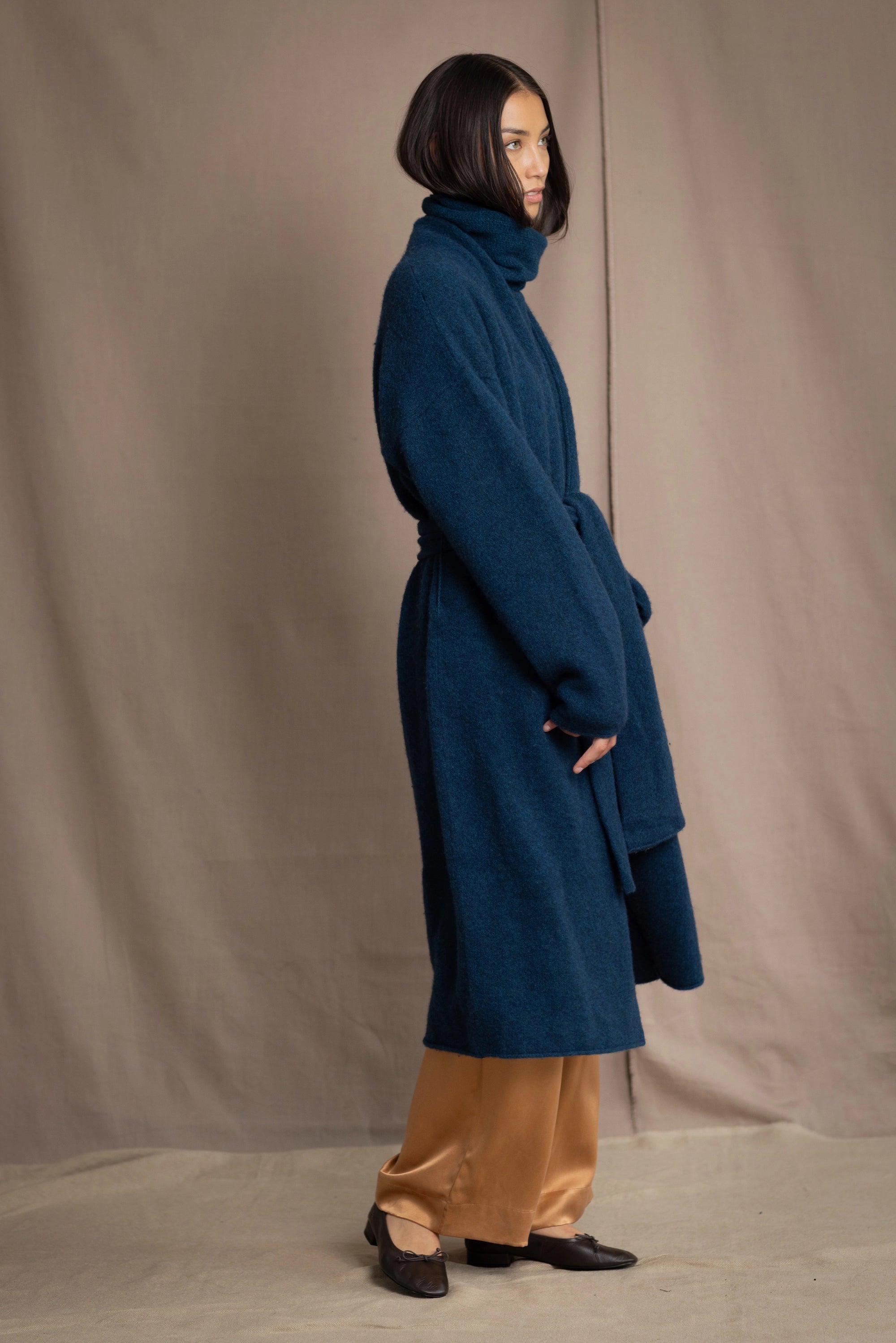 Woman wearing a long cashmere coat in teal blue over teal blue cashmere turtleneck sweater and bronze silk trousers