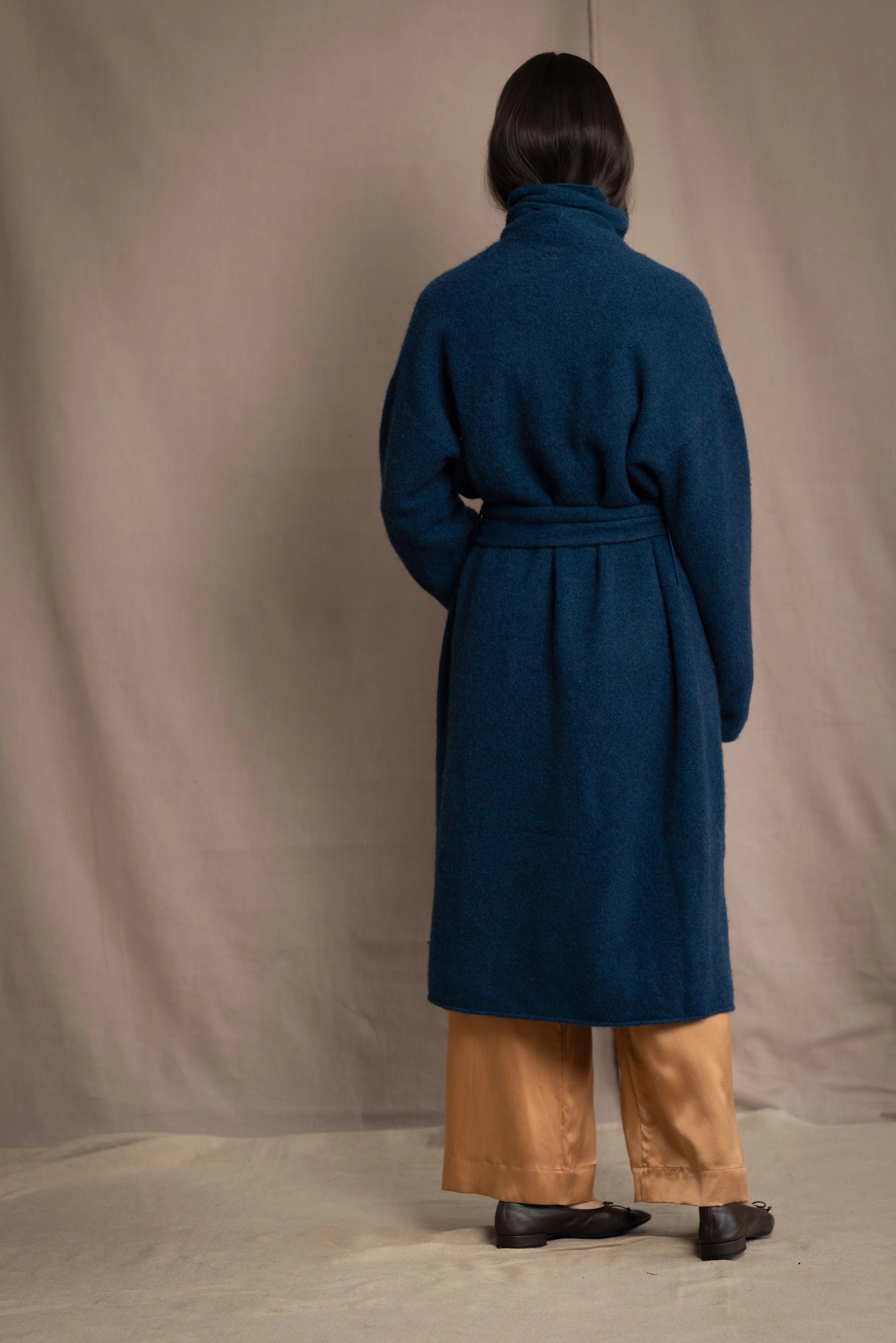 Woman wearing a long cashmere coat in teal blue over teal blue cashmere turtleneck sweater and bronze silk trousers