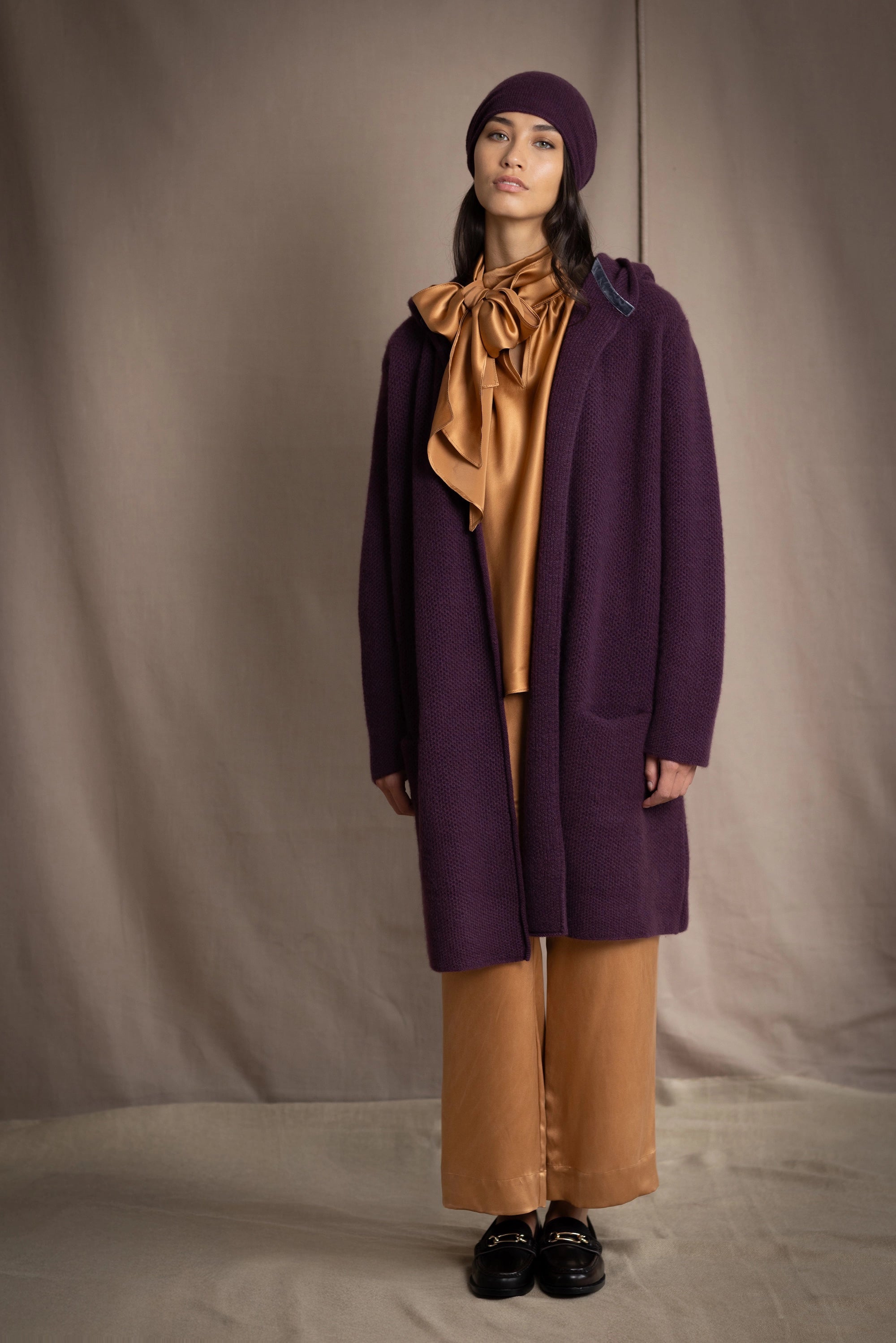 Woman wearing a long purple cashmere coat, bronze silk blouse and silk pants with dark purple cashmere beanie hat