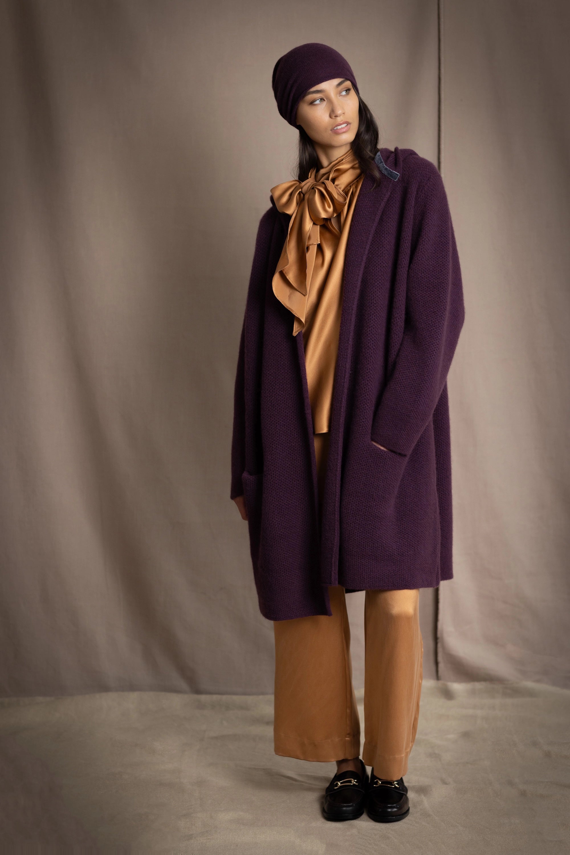 Woman wearing a long purple cashmere coat, bronze silk blouse and silk pants with dark purple cashmere beanie hat