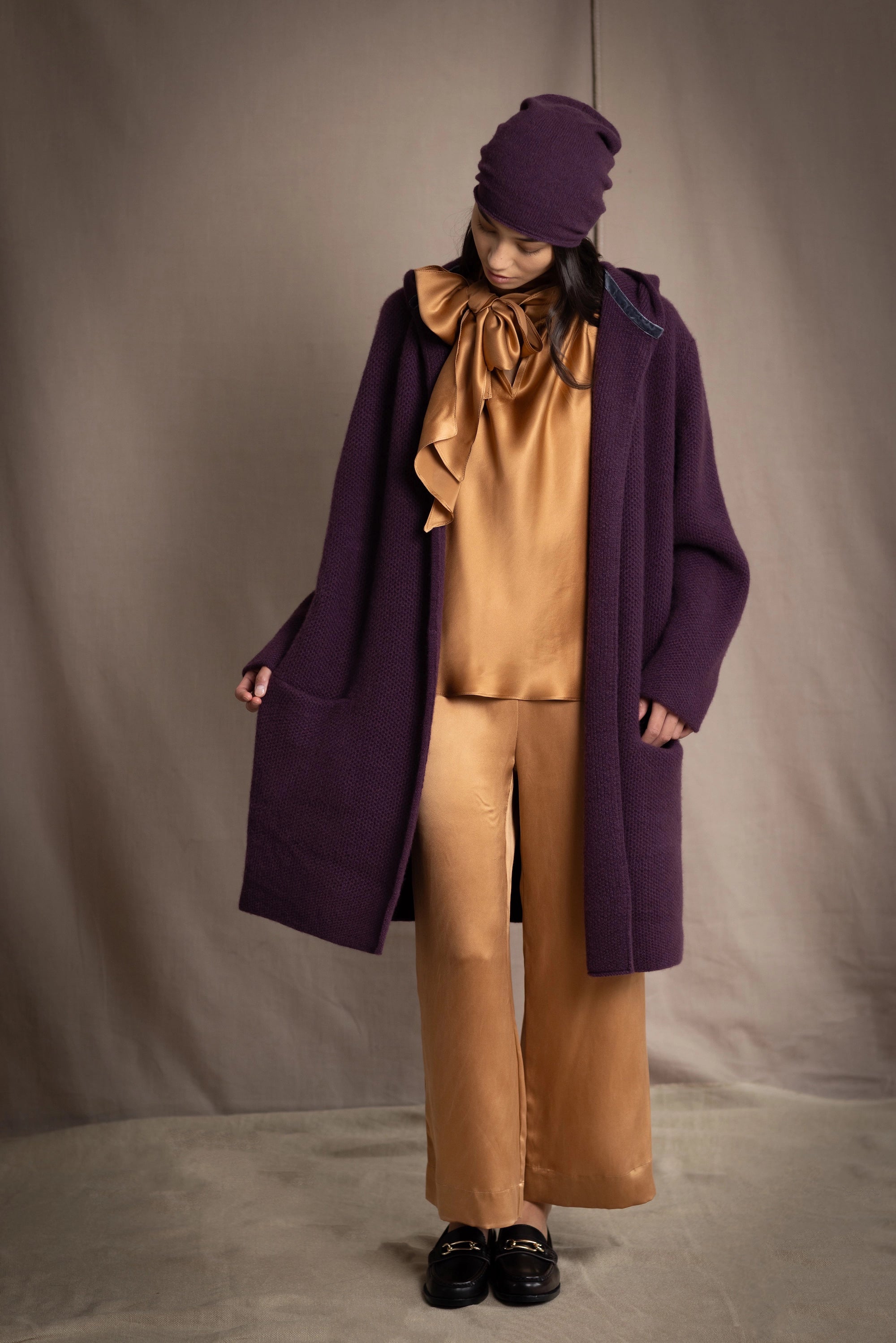 Woman wearing a long purple cashmere coat, bronze silk blouse and silk pants with dark purple cashmere beanie hat