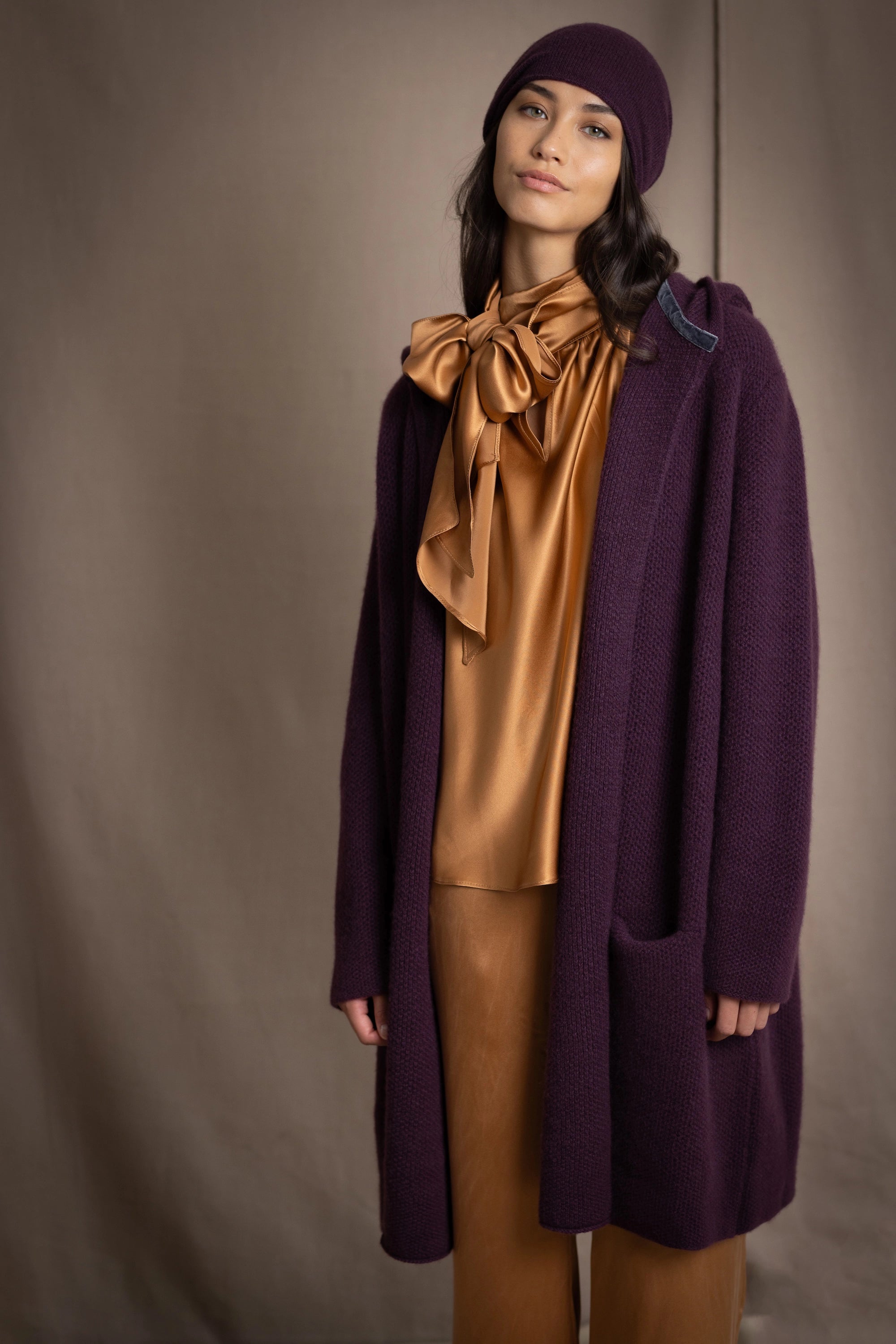Woman wearing a long purple cashmere coat, bronze silk blouse and silk pants with dark purple cashmere beanie hat