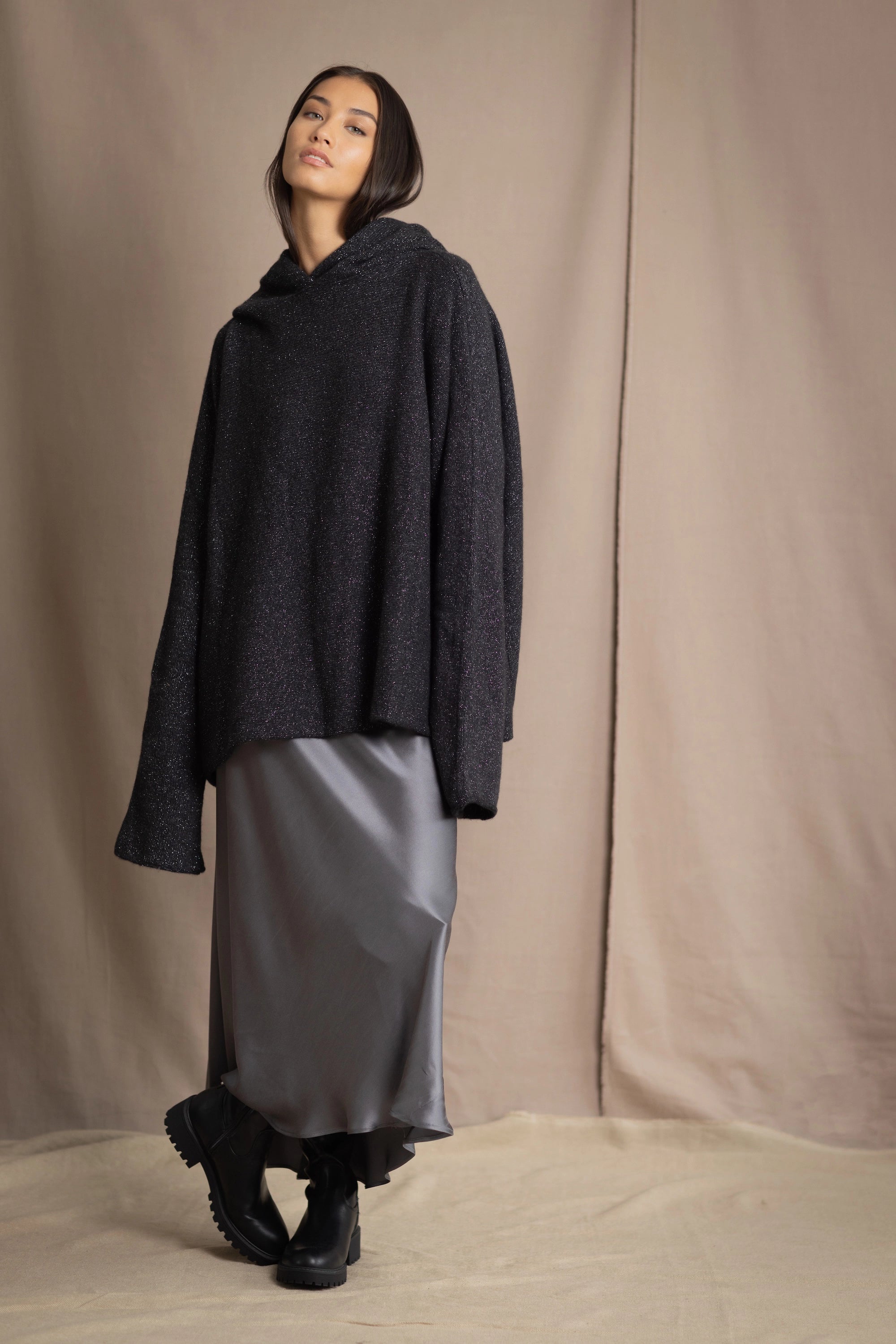 woman wearing an oversized cashmere sweater with hood in black and silver sparkly metallic colour with dark grey silk skirt and black leather boots