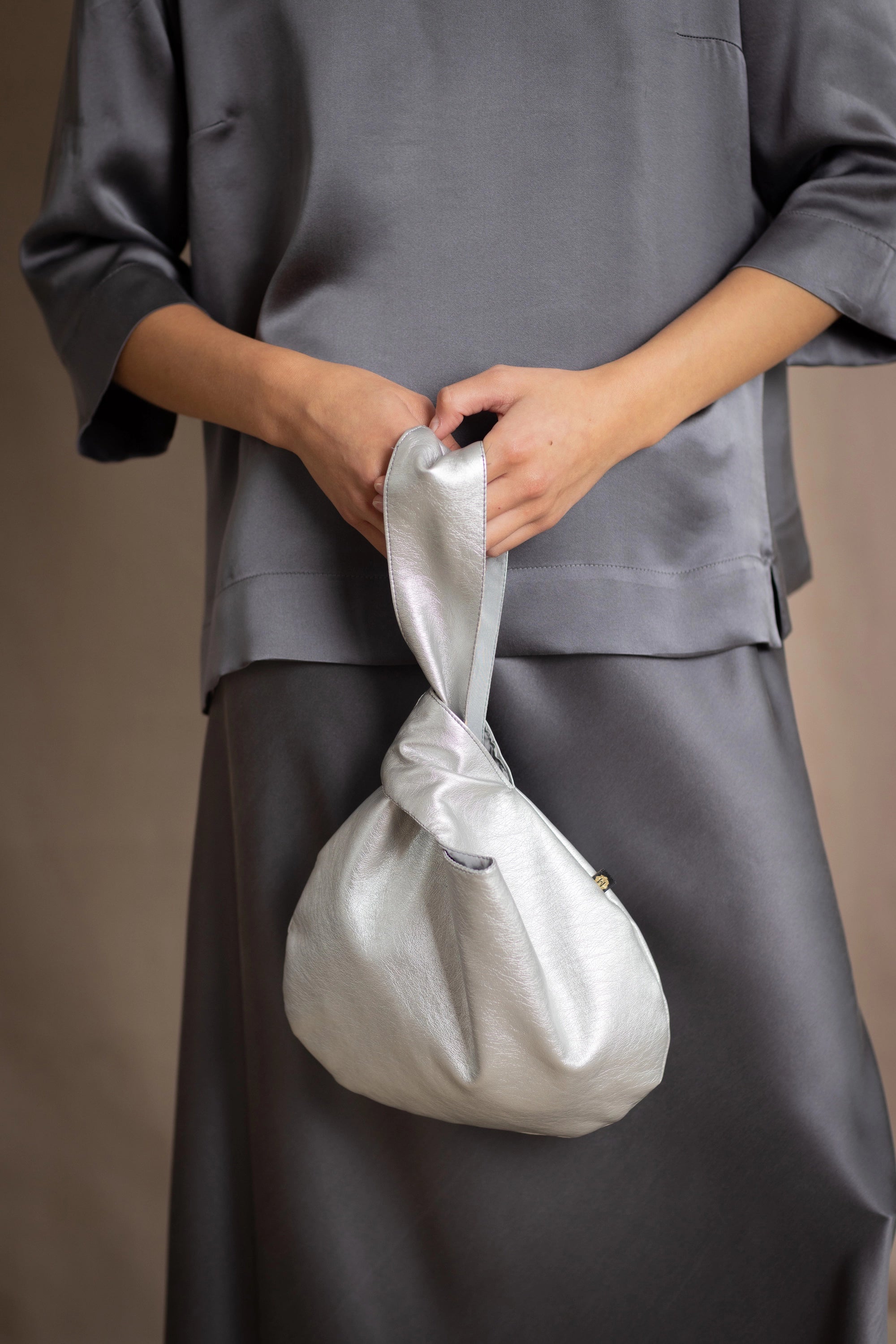 Silver vegan leather pull through handbag held by a person wearing a dark grey metallic silk outfit