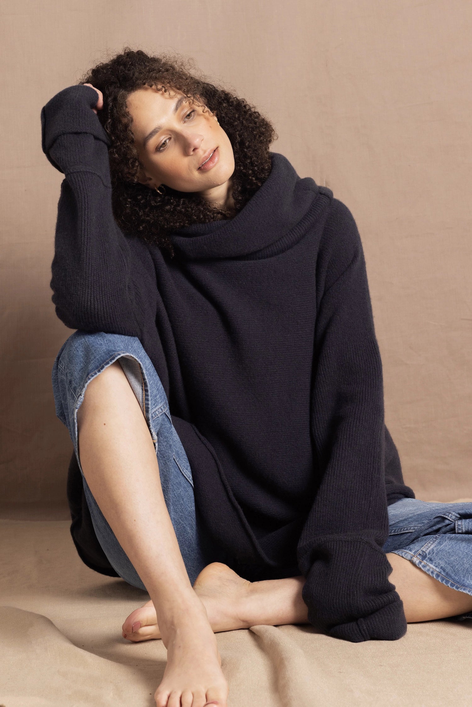 A woman sits wearing an oversized cashmere sweater in a grey blue colour with jeans