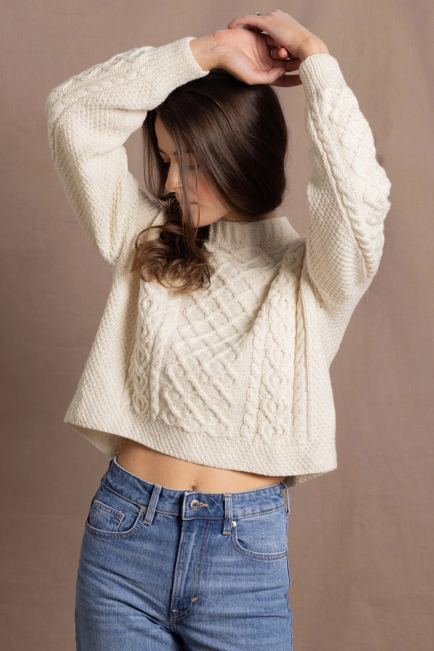 a woman wears a cropped cashmere aran sweater with her hands up in the air and wearing jeans