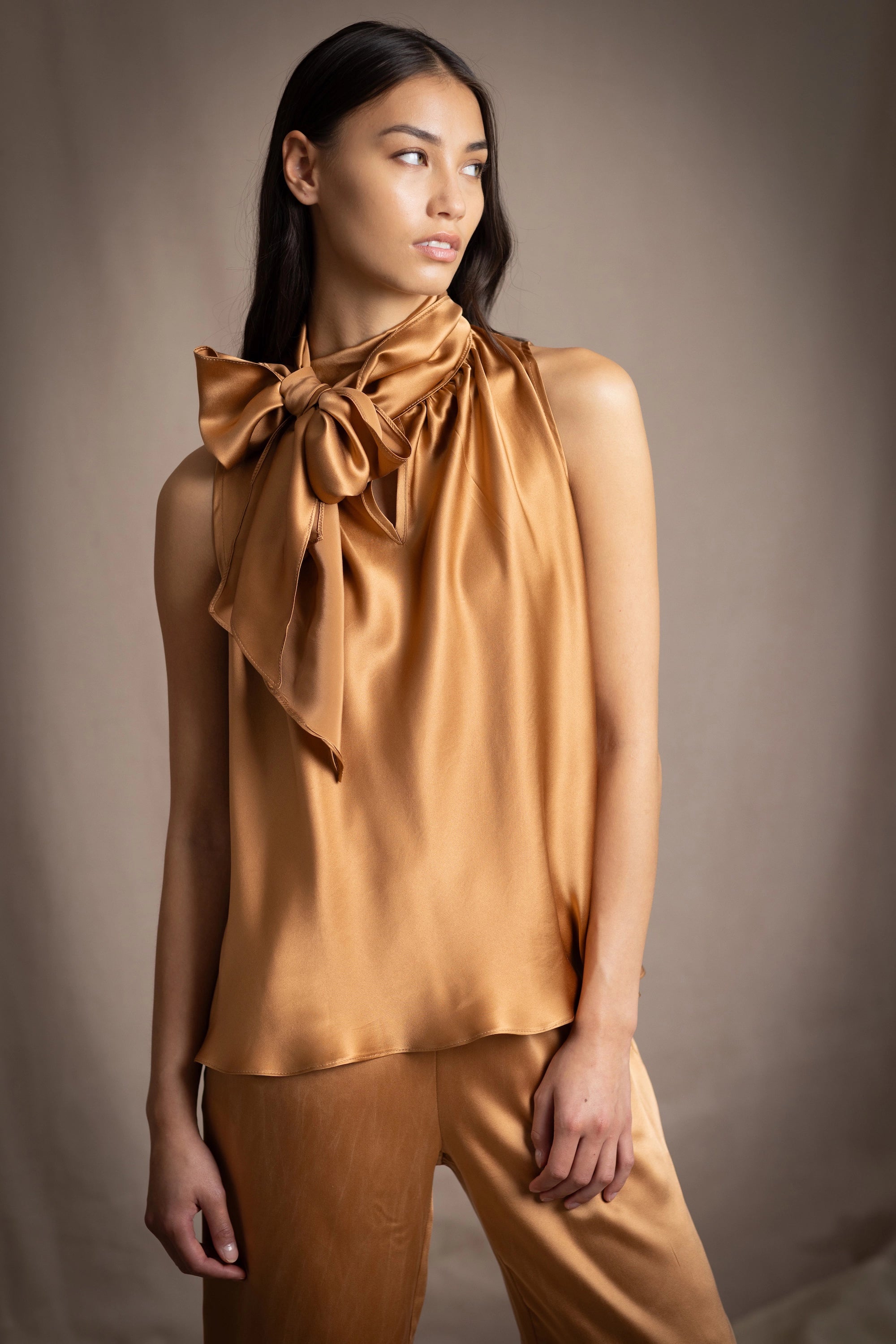 Woman wearing a bronze sleeveless silk pussy bow top with a large bow and matching pants