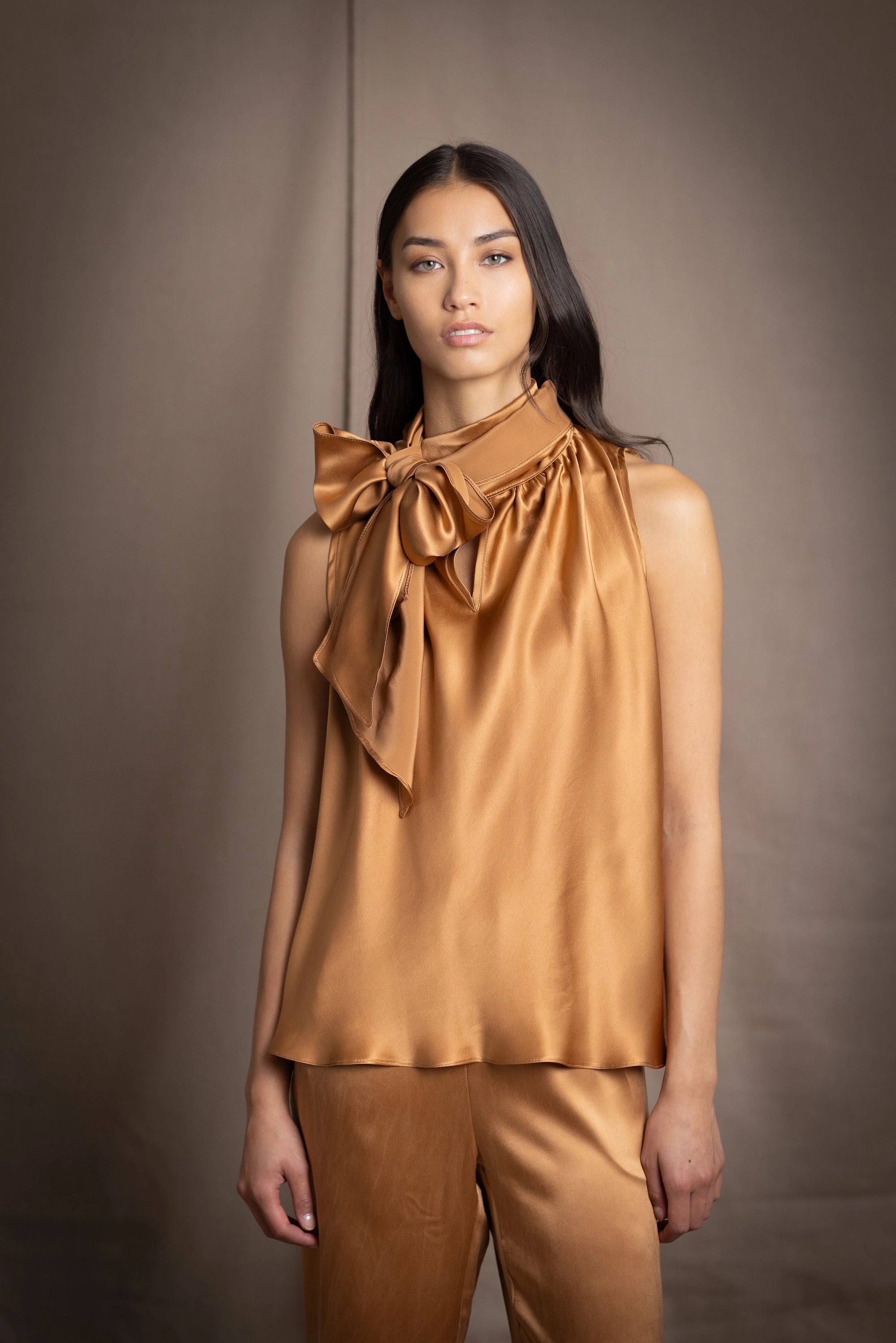 Woman wearing a bronze sleeveless silk pussy bow top with a large bow and matching pants